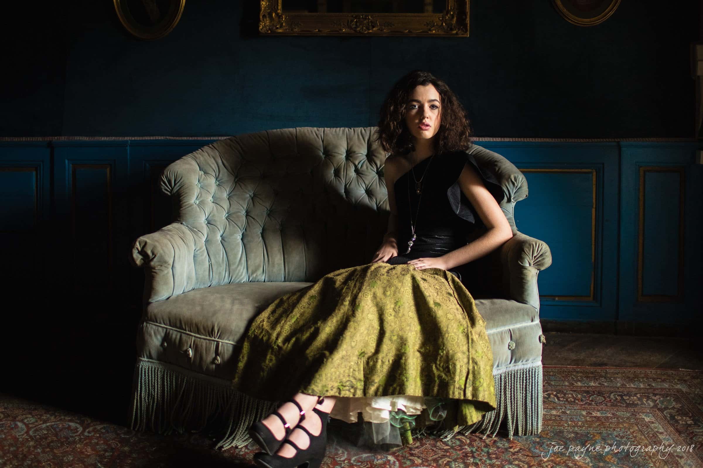 parker j pfister's french revolution workshop review A woman with curly hair sits on a vintage sofa in a dimly lit room with blue walls. She wears a black top and a long olive skirt, with black strappy shoes. A large mirror hangs on the wall behind her. The atmosphere is calm and introspective.