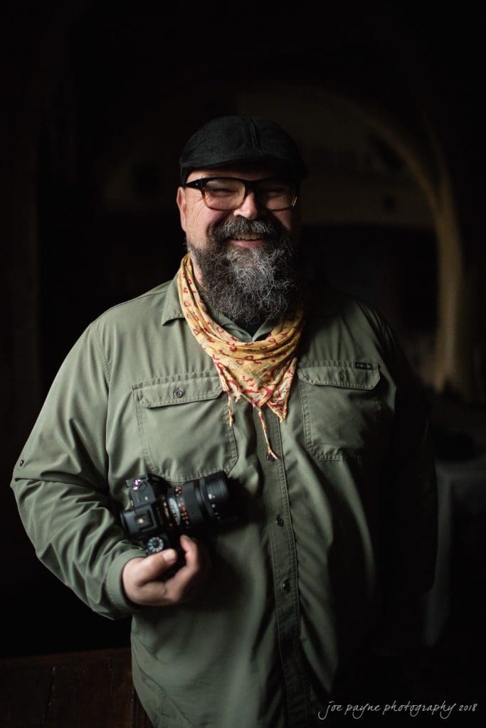 parker j pfister's french revolution workshop review A bearded man wearing a black cap, green shirt, and floral scarf stands smiling with a camera in hand. The background is dimly lit.