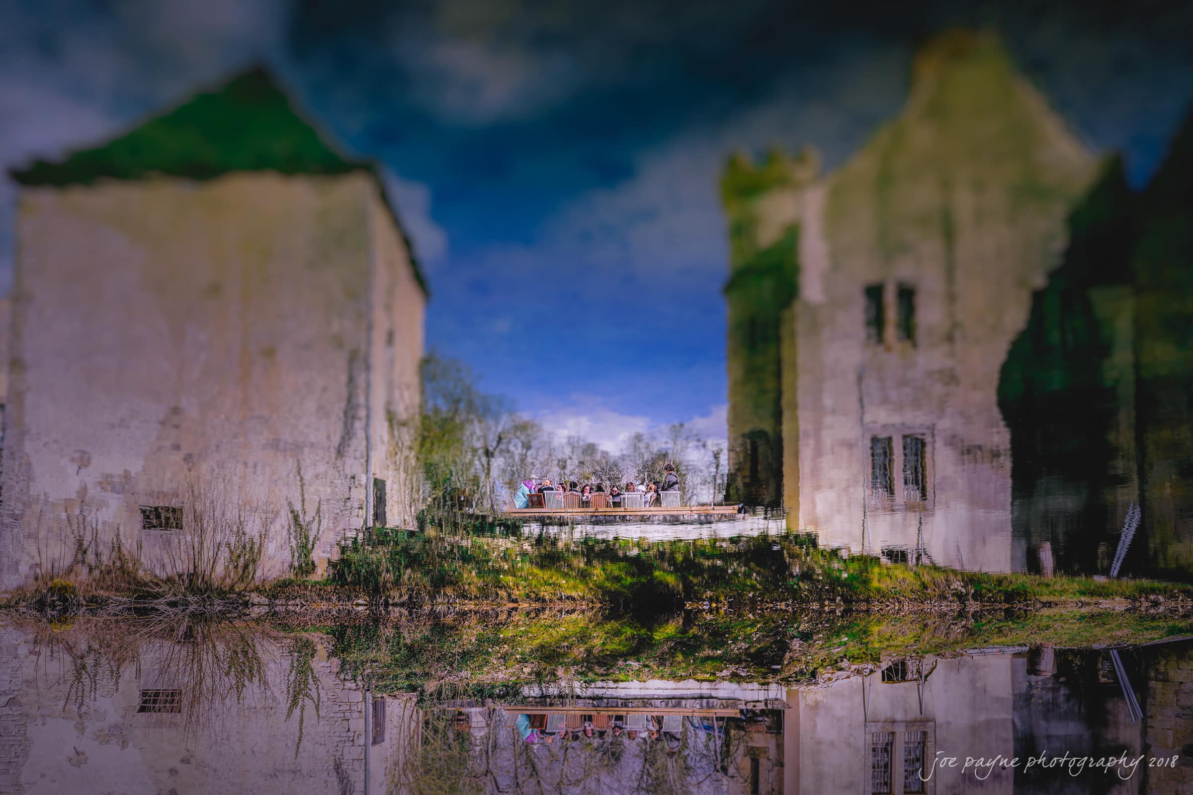 parker j pfister's french revolution workshop review A surreal reflection of two old, weathered buildings in a body of water. The scene includes a group of people gathered on a small platform between the structures, surrounded by trees and grass under a clear blue sky.