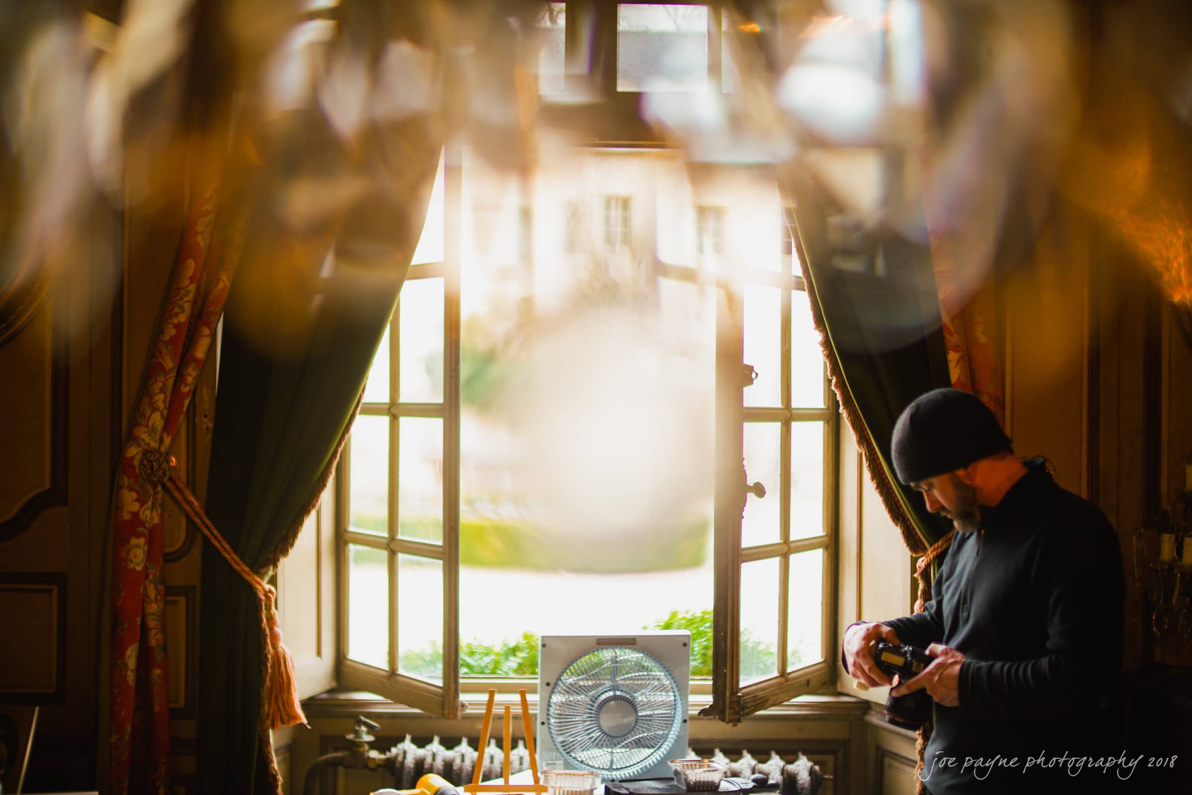 parker j pfister's french revolution workshop review A person in a dark beanie stands near a large, ornate window with curtains, inspecting a camera. The foreground is blurred, adding a dreamy effect to the cozy interior scene.