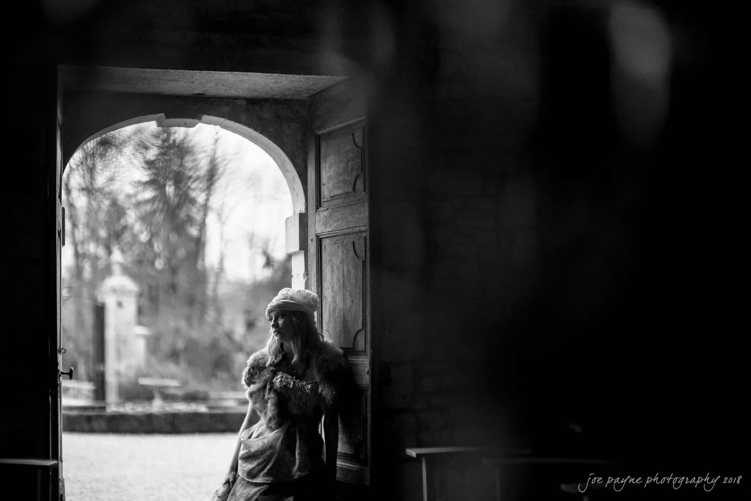 parker j pfister's french revolution workshop review A person dressed in historical clothing stands thoughtfully at the entrance of a stone building, illuminated by soft natural light. The open door reveals a blurred garden scene outside. The image is in black and white, adding a timeless feel.