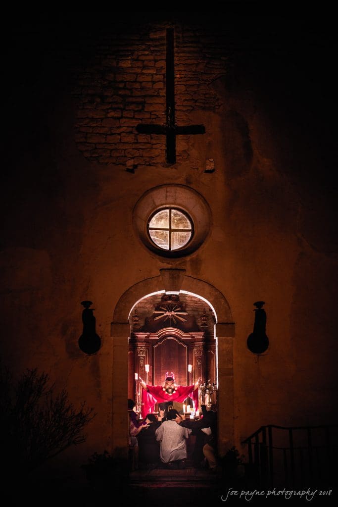 parker j pfister's french revolution workshop review A dimly lit church with a large cross above the entrance. Inside, people are gathered in front of an ornately decorated altar, illuminated by soft, warm light. The atmosphere is solemn and reverent.