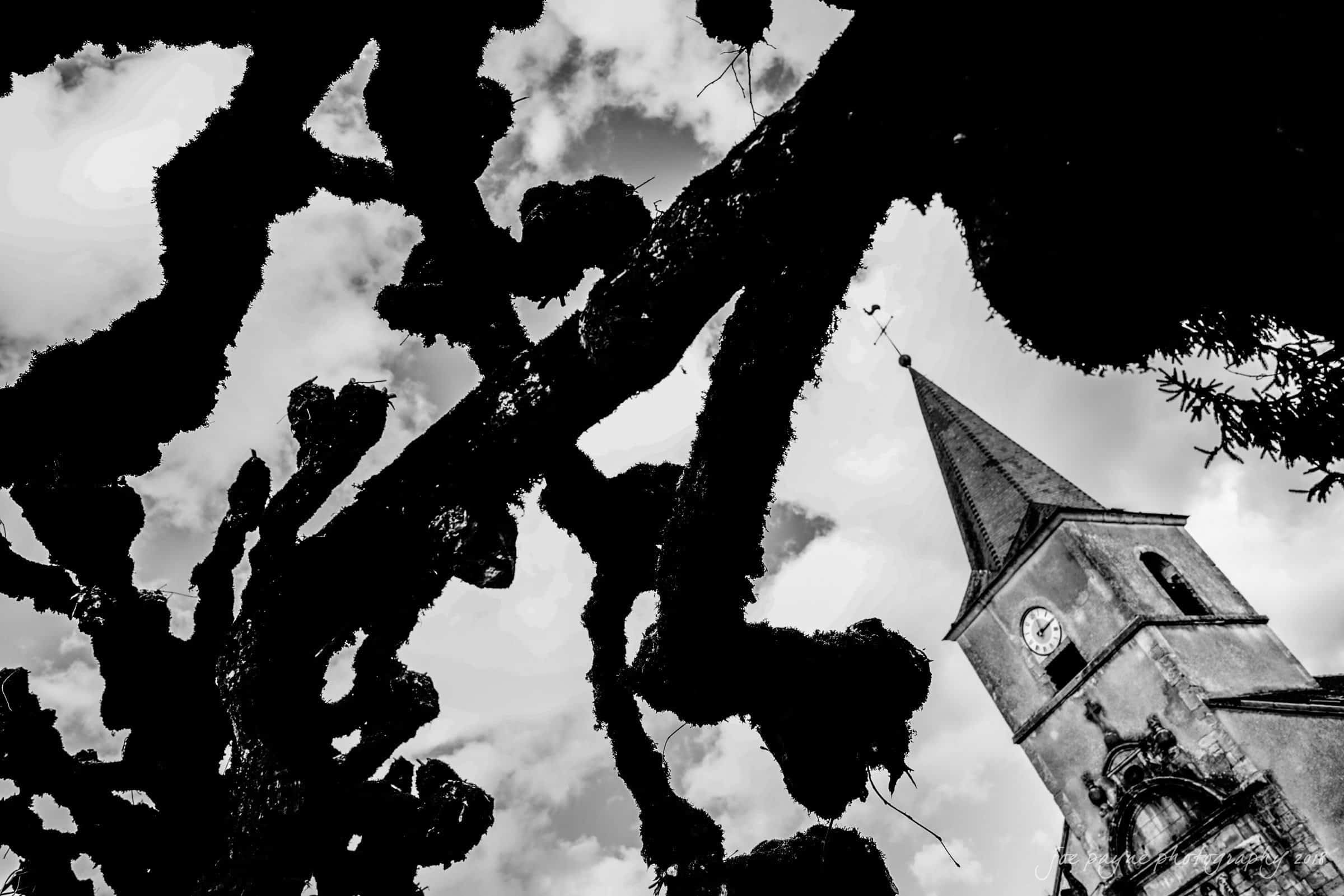 parker j pfister's french revolution workshop review A black-and-white image featuring silhouetted tree branches in the foreground and a church tower with a clock in the background against a cloudy sky. The dramatic contrast highlights the architectural details and natural forms.
