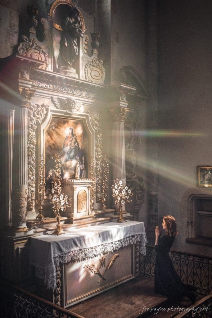 parker j pfister's french revolution workshop review A person kneeling in prayer in front of an ornate altar with religious imagery. Sunlight streams through a nearby window, casting light onto the scene. The altar is adorned with floral decorations and intricately carved details.