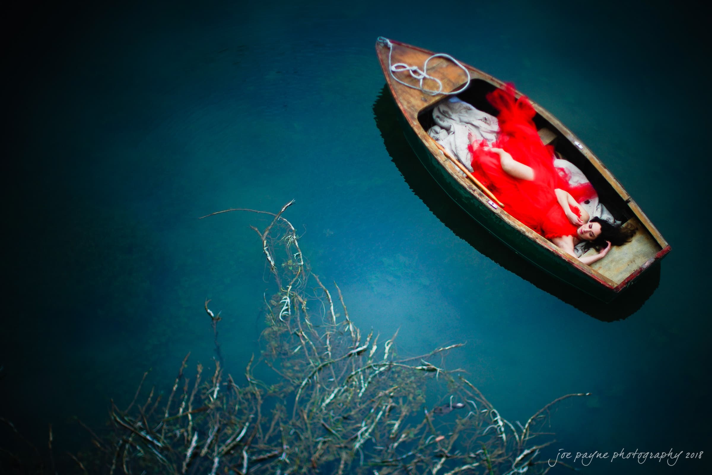 In a scene reminiscent of Parker J. Pfister's ethereal style, a woman in a flowing red dress lies in a small wooden boat on a calm, blue-green lake. Bare branches extend from the bottom left, crafting a serene and dreamy tableau that could fit right into his French Revolution workshop.