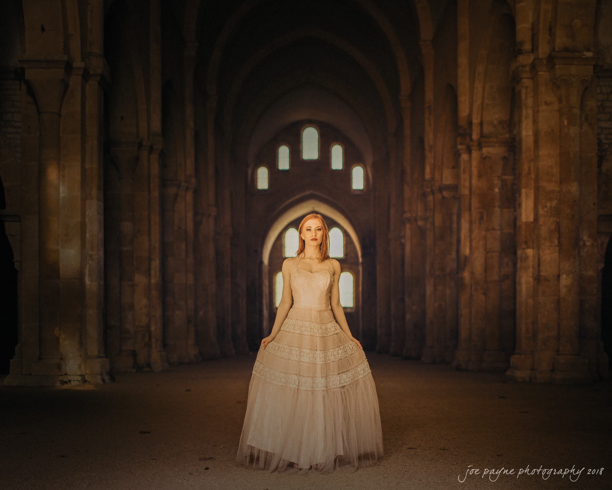 parker j pfister's french revolution workshop review A woman stands in the center of a dimly lit, arched hallway, wearing a flowing, strapless gown. Warm light filters in from a series of windows, casting a serene ambiance.