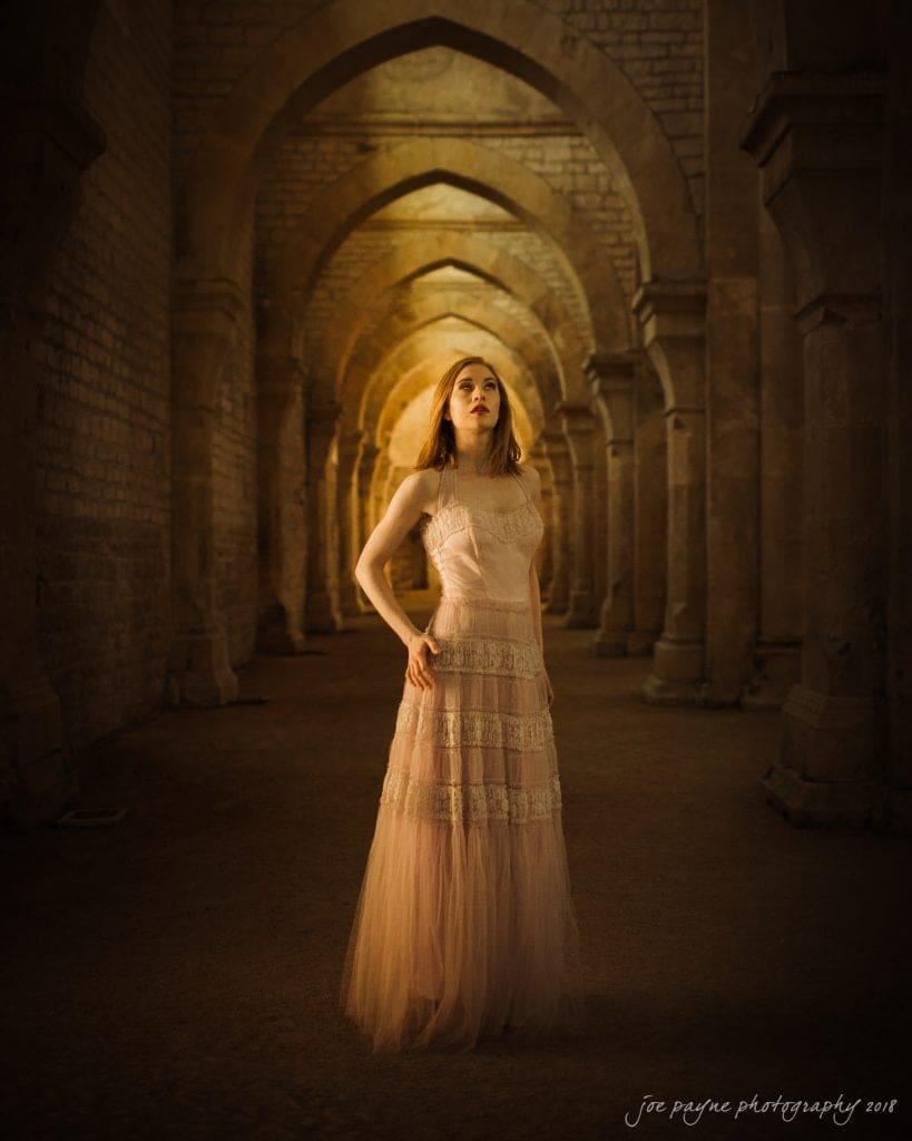 parker j pfister's french revolution workshop review A woman in a long, elegant pink dress stands in a dimly lit stone archway. She gazes upward with a serene expression, surrounded by a series of symmetrical arches creating a dramatic, atmospheric setting.