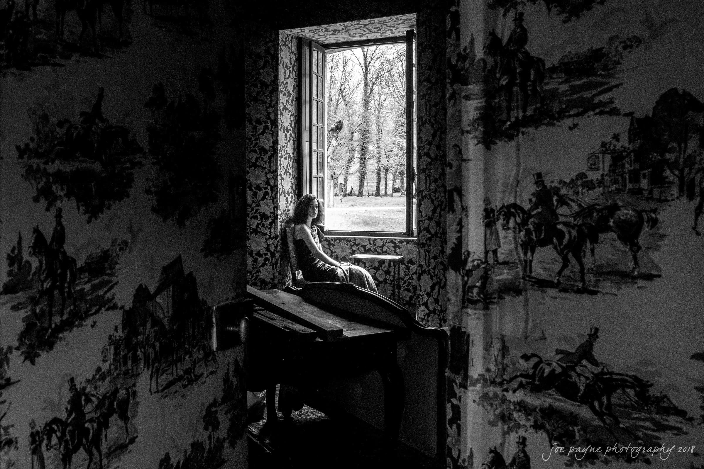 parker j pfister's french revolution workshop review A black and white photo shows a person sitting on a windowsill, gazing outside. The room is dimly lit with patterned wallpaper depicting scenes with people and horses. Light from the window illuminates the room, contrasting with the indoor shadows.