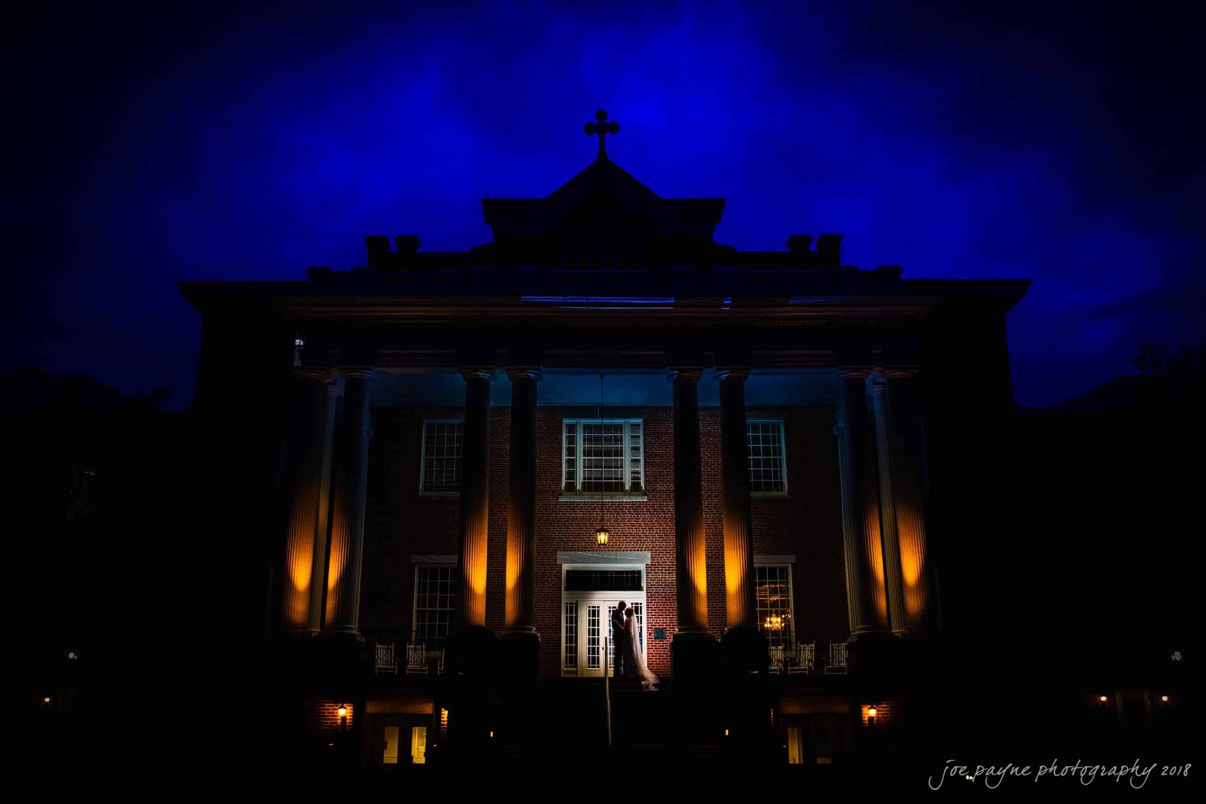st. mary's & umstead wedding photographer - sarah & jake A large brick building with pillars is dramatically lit at night. The sky is deep blue, and the building casts a warm glow from lights below. A silhouette of two people is visible near the entrance.