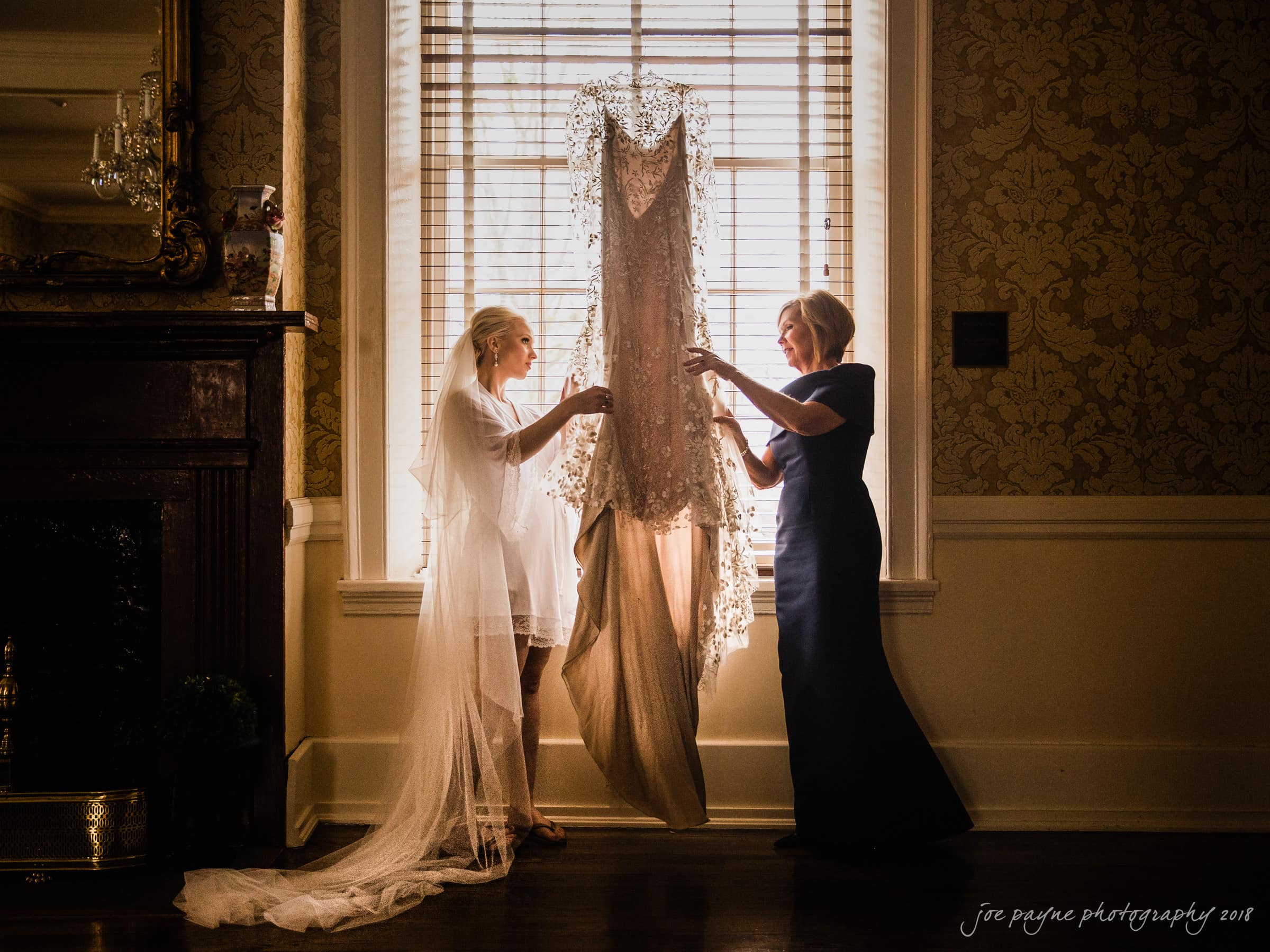 st. mary's & umstead wedding photographer - sarah & jake Two women stand in a warmly lit room, holding a lace wedding dress in front of a window. One wears a bridal robe, the other a dark gown. The scene conveys a sense of anticipation and intimacy.