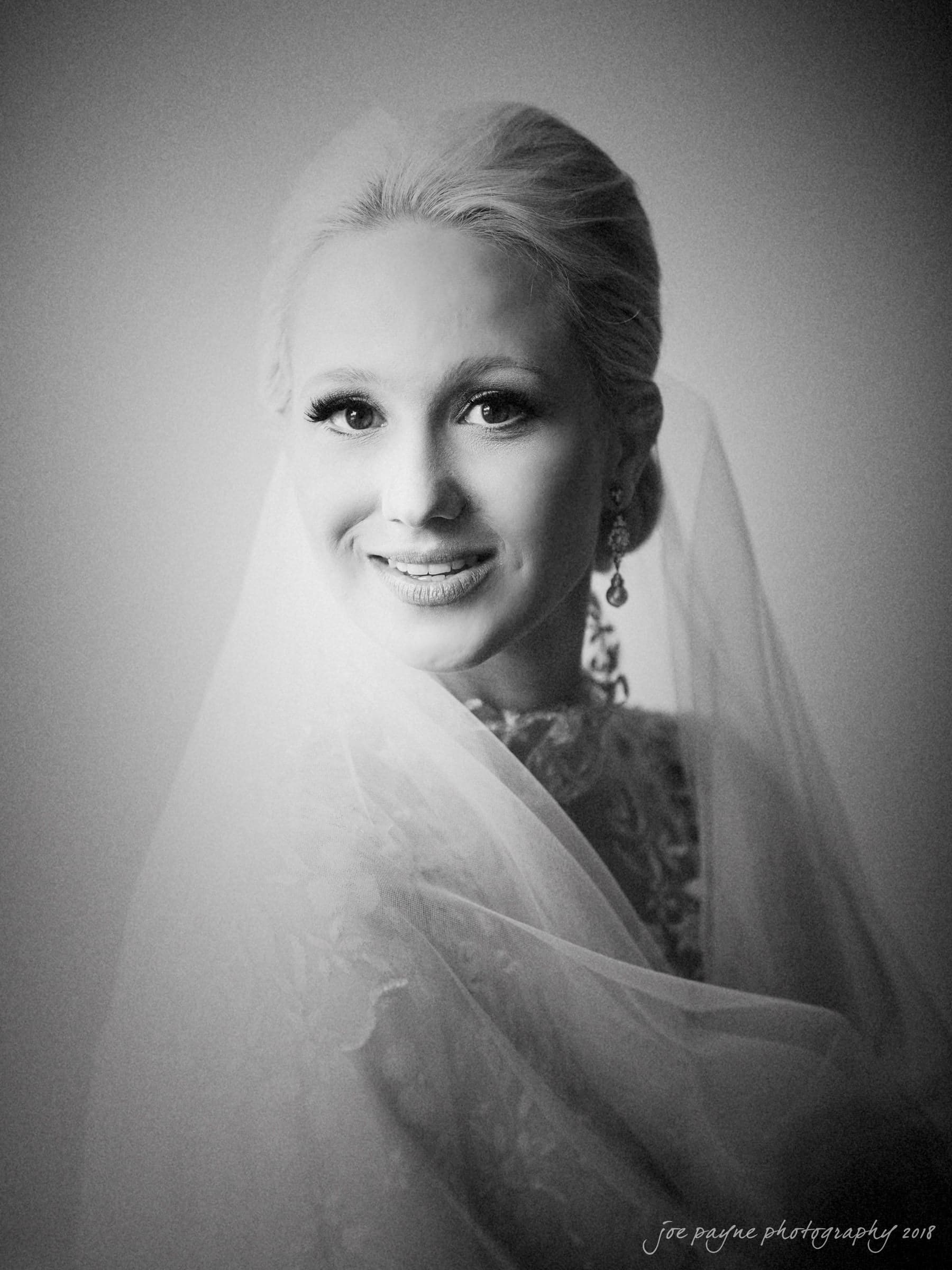 st. mary's & umstead wedding photographer - sarah & jake Black and white portrait of a bride smiling. She is wearing a lace wedding dress and veil, with earrings visible. The soft lighting creates a gentle and serene atmosphere.