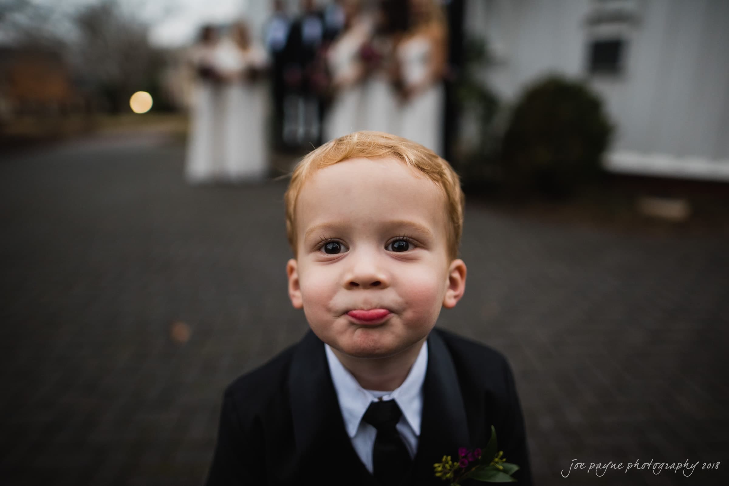 st. mary's & umstead wedding photographer - sarah & jake Young boy with light brown hair in a black suit stands in focus, playfully pursing his lips. In the blurred background, people dressed in formal attire are gathered, suggesting an event. The setting is outdoors with stone paving.