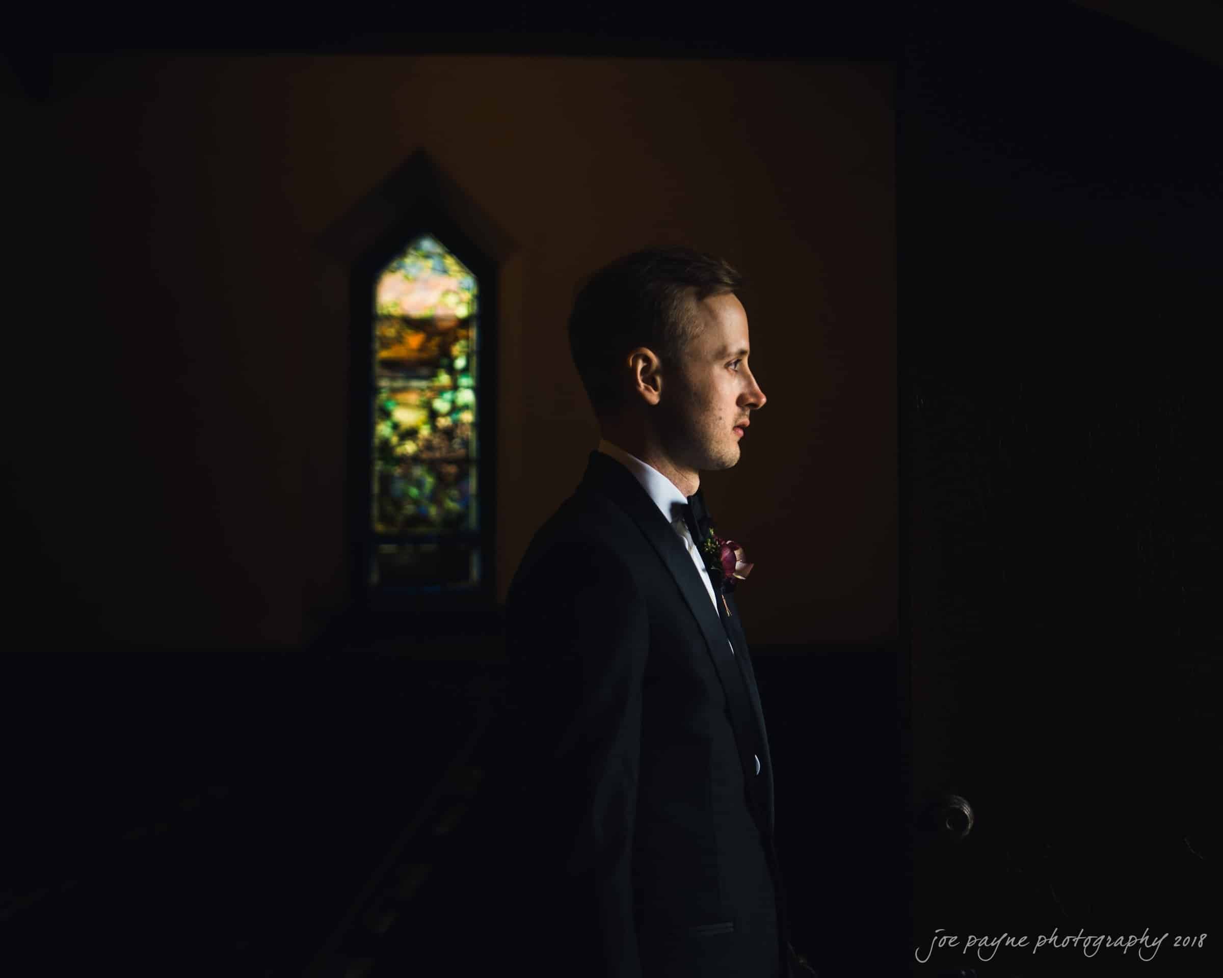 st. mary's & umstead wedding photographer - sarah & jake Man in a suit standing in a dimly lit room with stained glass window in the background, light highlighting his face.