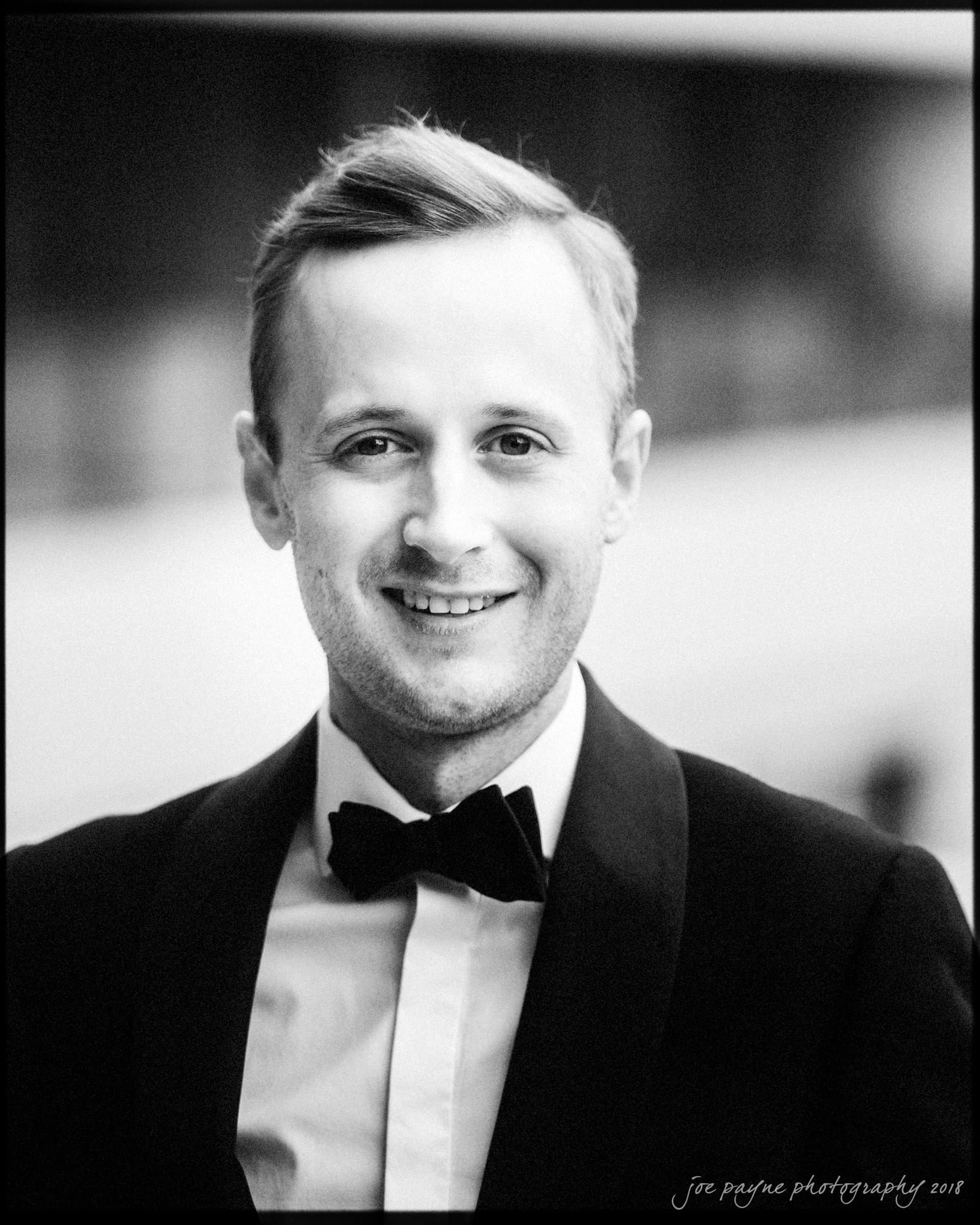 st. mary's & umstead wedding photographer - sarah & jake A man smiling while wearing a tuxedo and bow tie in a black and white photograph.