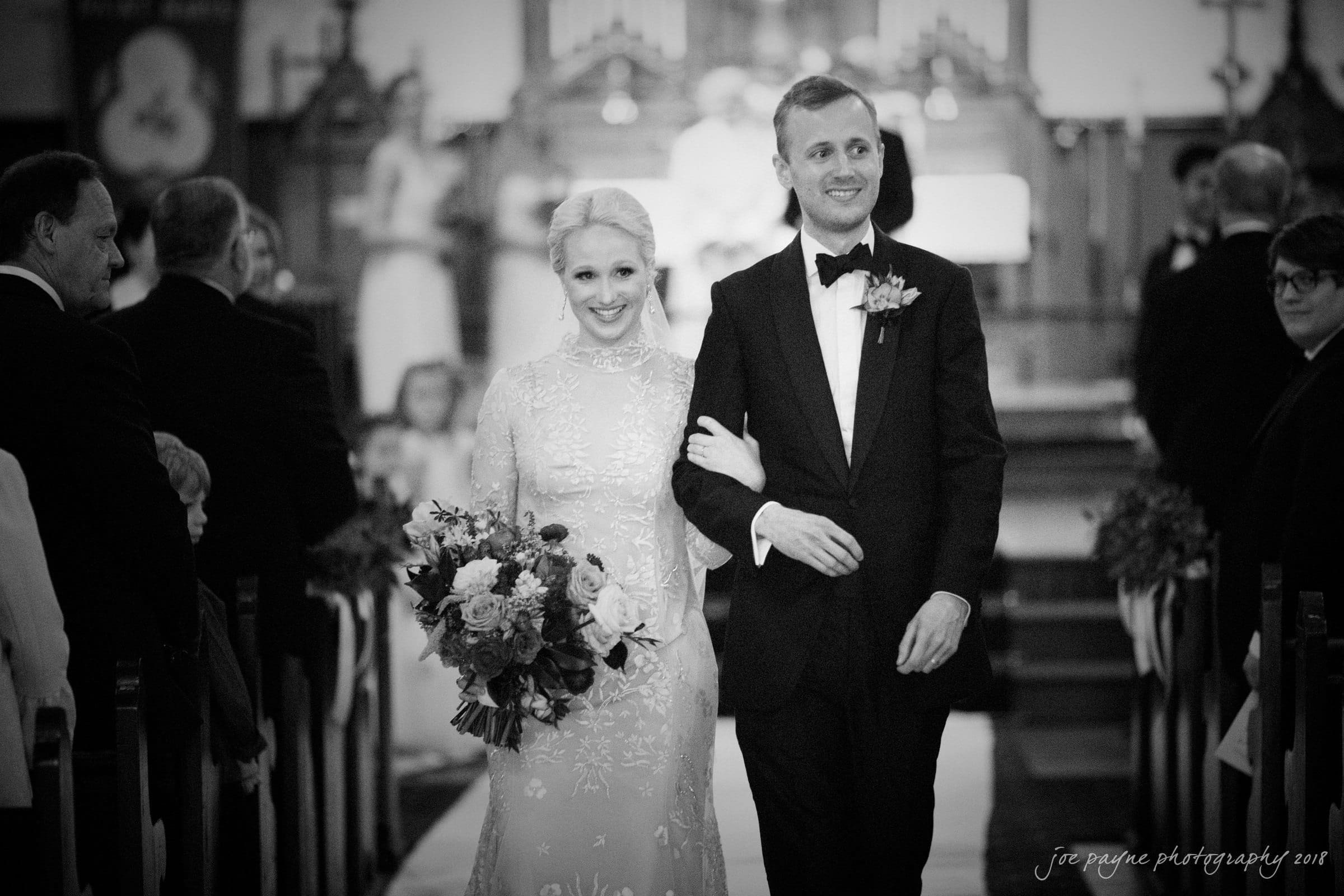 st. mary's & umstead wedding photographer - sarah & jake A bride and groom walk down the aisle arm in arm. The groom wears a black tuxedo, and the bride holds a bouquet of flowers, wearing a white lace dress. People are seated on either side, and the background shows a decorated altar.