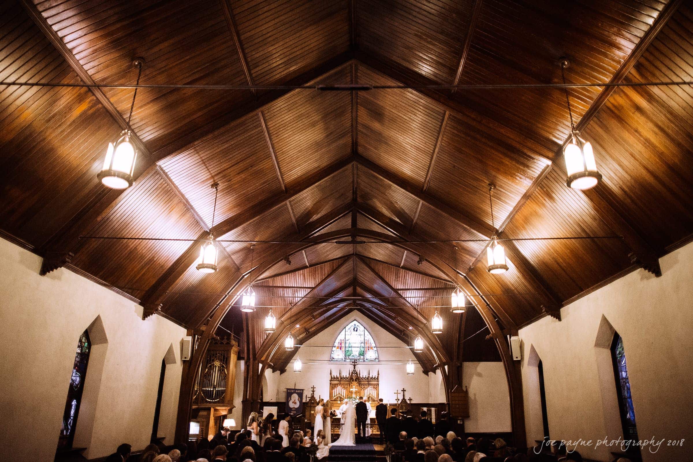 st. mary's & umstead wedding photographer - sarah & jake A warmly-lit church interior with a wooden vaulted ceiling. Rows of pews are filled with people witnessing a ceremony at the altar. Stained glass windows and hanging lights add to the serene atmosphere.