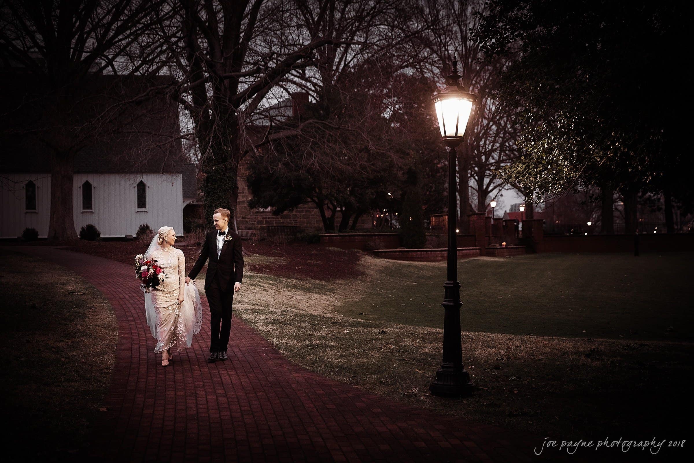 A bride and groom walk hand in hand along a brick path at dusk, surrounded by trees and illuminated by a streetlamp. Captured beautifully by St. Marys Umstead wedding photographer, the bride holds a bouquet as they both smile warmly at each other.
