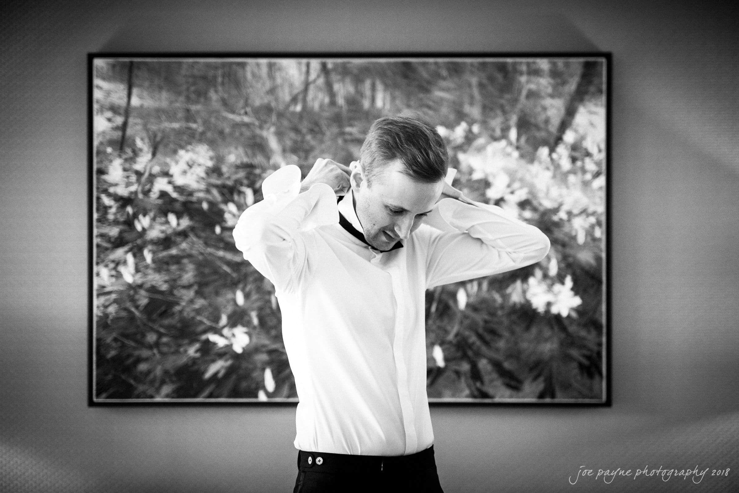 st. mary's & umstead wedding photographer - sarah & jake A black and white photo of a person adjusting their shirt collar in front of a large painting depicting flowers. The persons expression is focused, and the setting appears to be indoors.