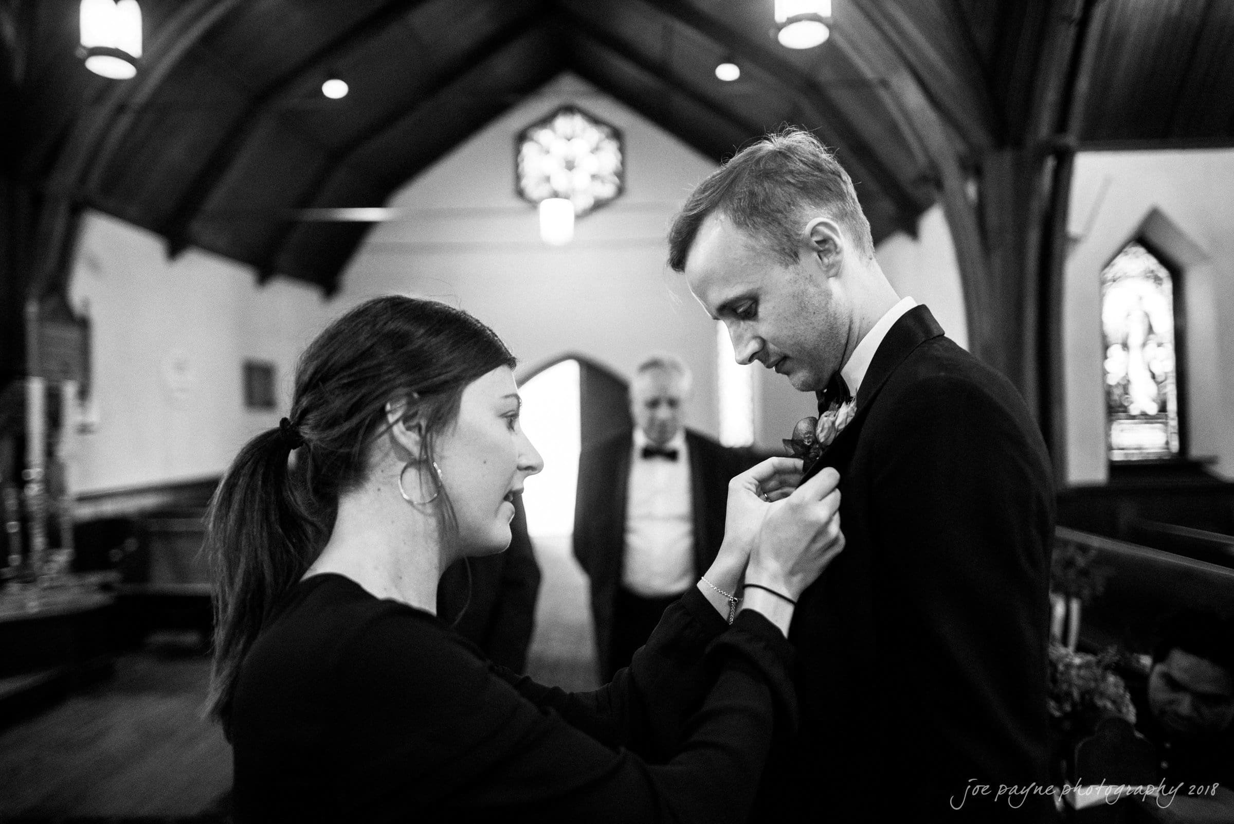 st. mary's & umstead wedding photographer - sarah & jake A woman adjusts a boutonniere on a mans suit inside a church. Another man in the background, wearing a tuxedo, looks on. The setting is dimly lit with visible stained glass windows and wooden beams.