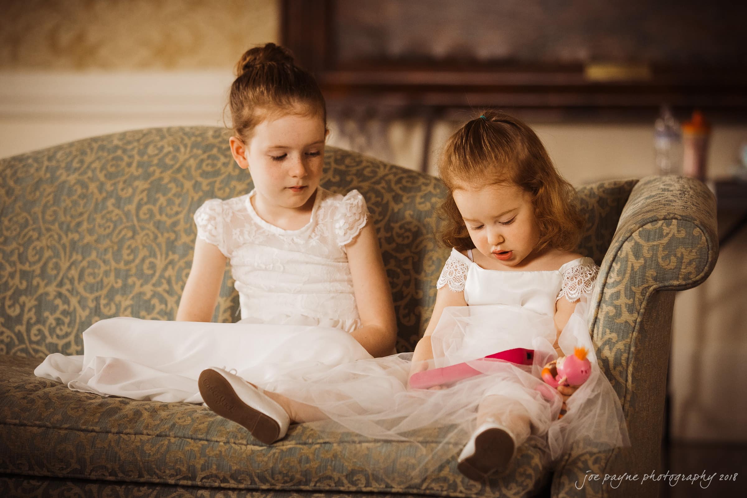 st. mary's & umstead wedding photographer - sarah & jake Two young girls in white dresses sit on a patterned sofa. One girl attentively looks at a pink toy in the others lap. The room has warm lighting and a classic, elegant decor.