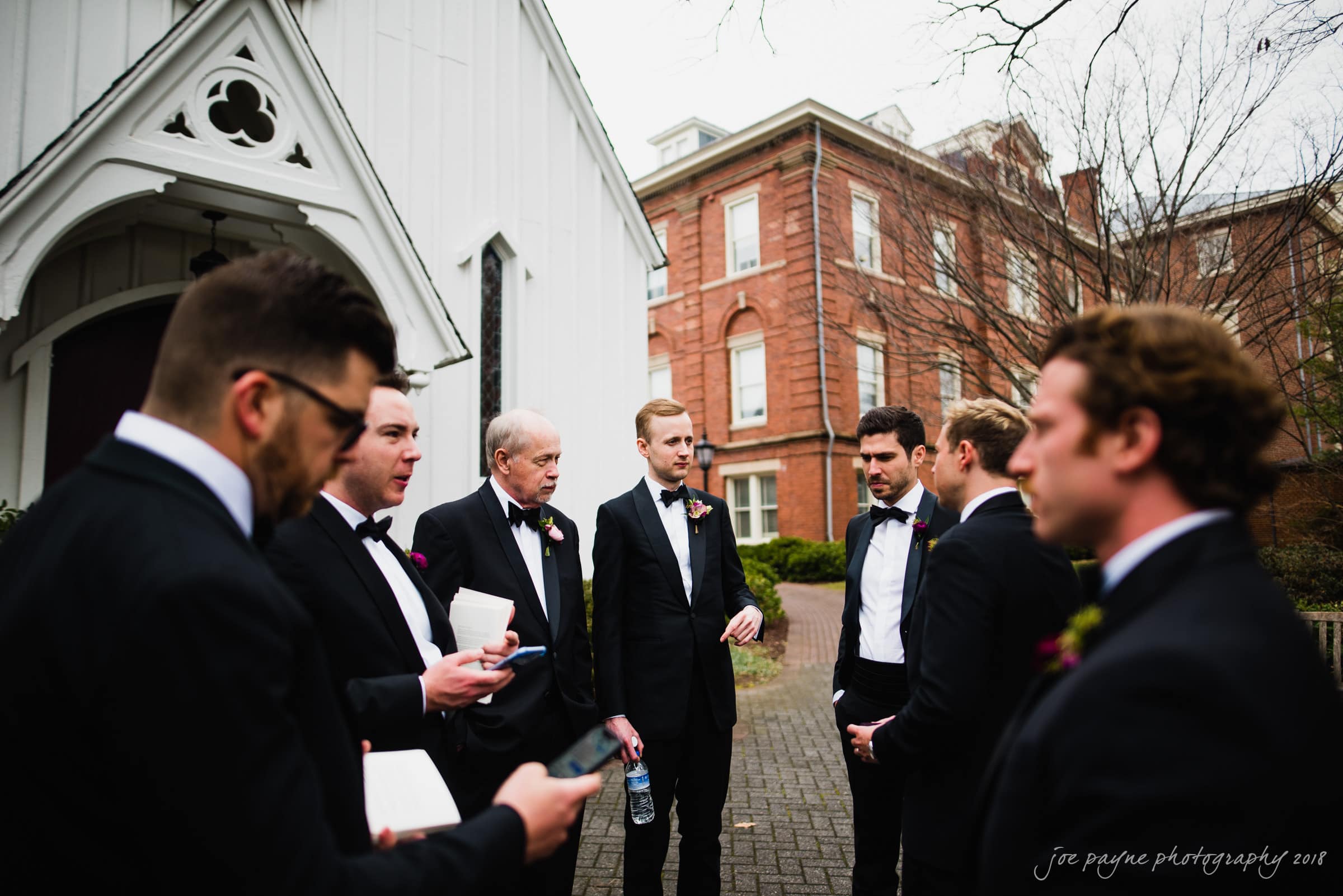 st. mary's & umstead wedding photographer - sarah & jake A group of men in suits stand outside a white church building, engaging in conversation. Some hold papers and a water bottle, while trees and a brick building are visible in the background.