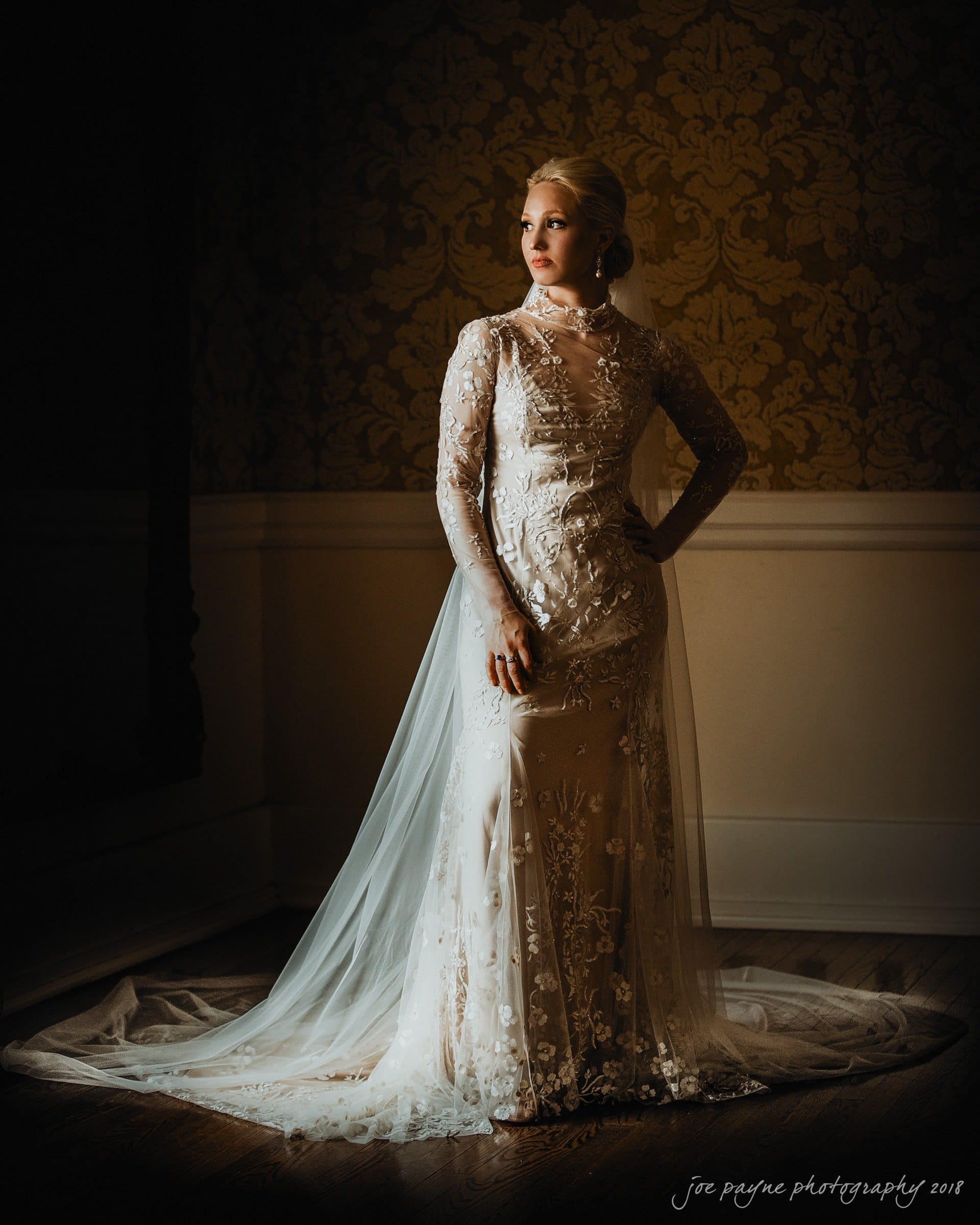 st. mary's & umstead wedding photographer - sarah & jake A woman in an elegant, embroidered white wedding dress stands in a softly lit room with ornate wallpaper. She gazes to the side with one hand on her hip, and a long veil trails behind her, illuminated by the light.