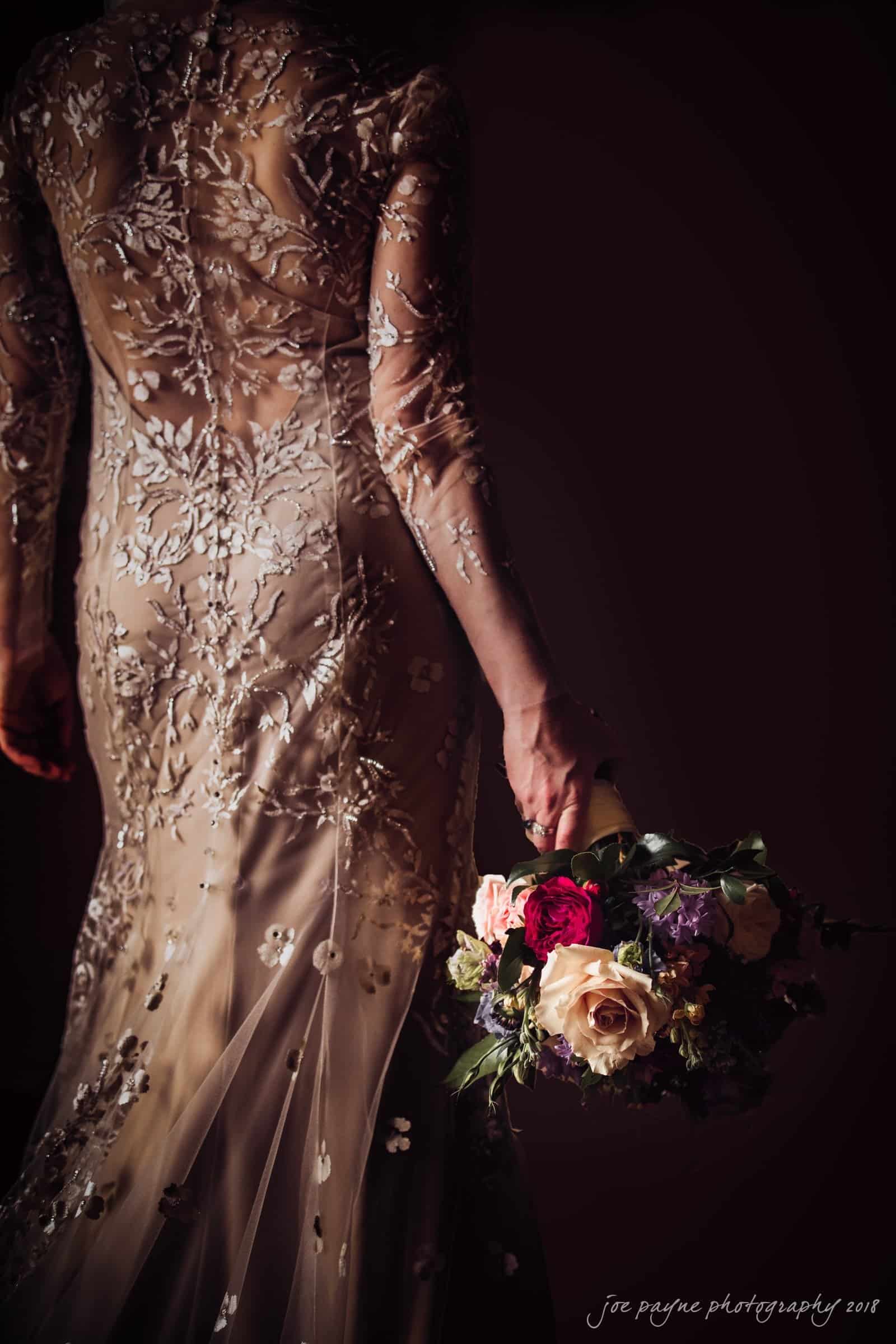 st. mary's & umstead wedding photographer - sarah & jake A person in an elegant, floral-embroidered dress holds a bouquet with roses and other flowers. The background is dark, highlighting the intricate details of the attire and the colorful blooms.
