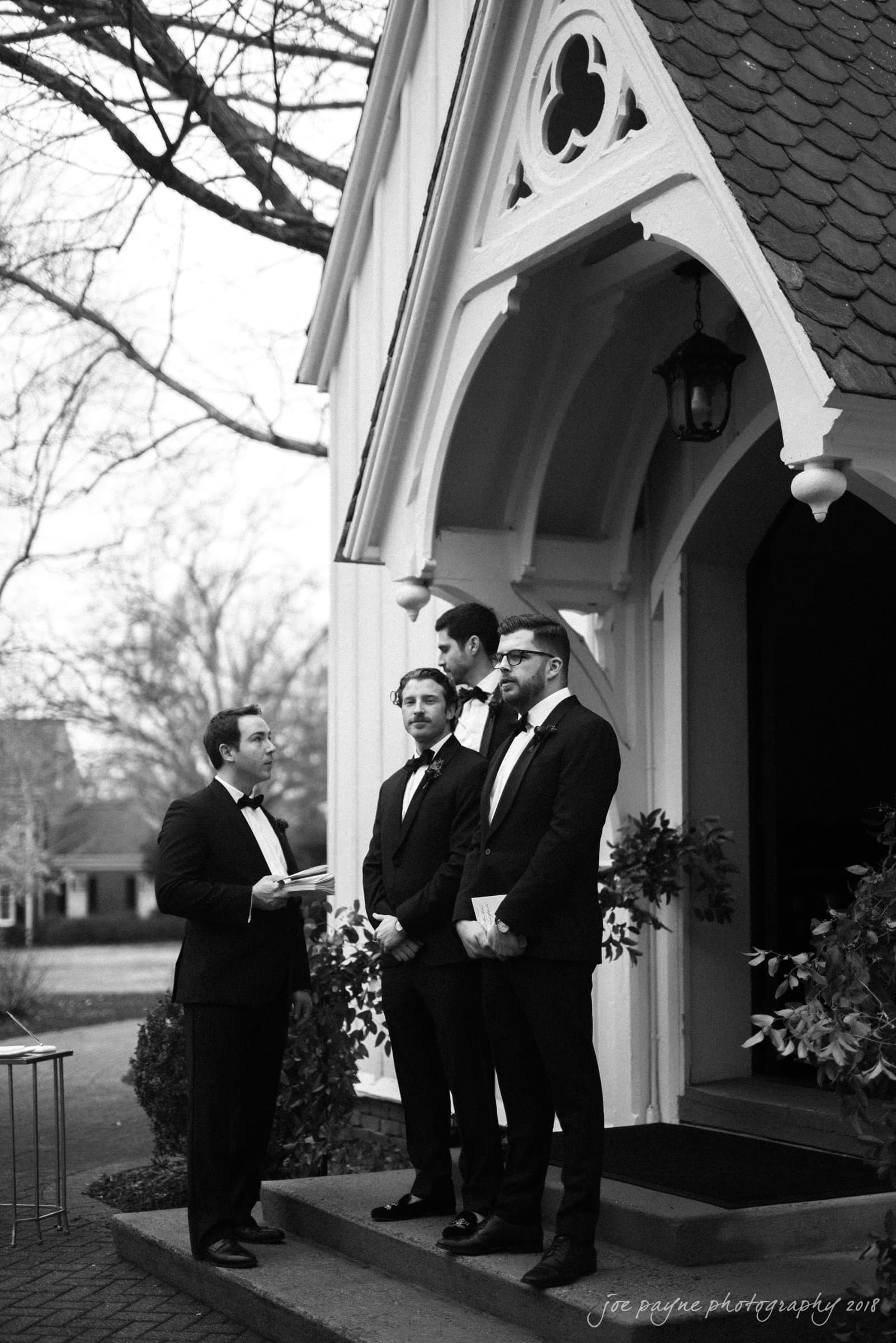 st. mary's & umstead wedding photographer - sarah & jake Four men in formal suits stand outside a small chapel, engaged in conversation. The chapel has a pointed arch and a shingled roof. Trees with bare branches are visible in the background, suggesting a cool season.