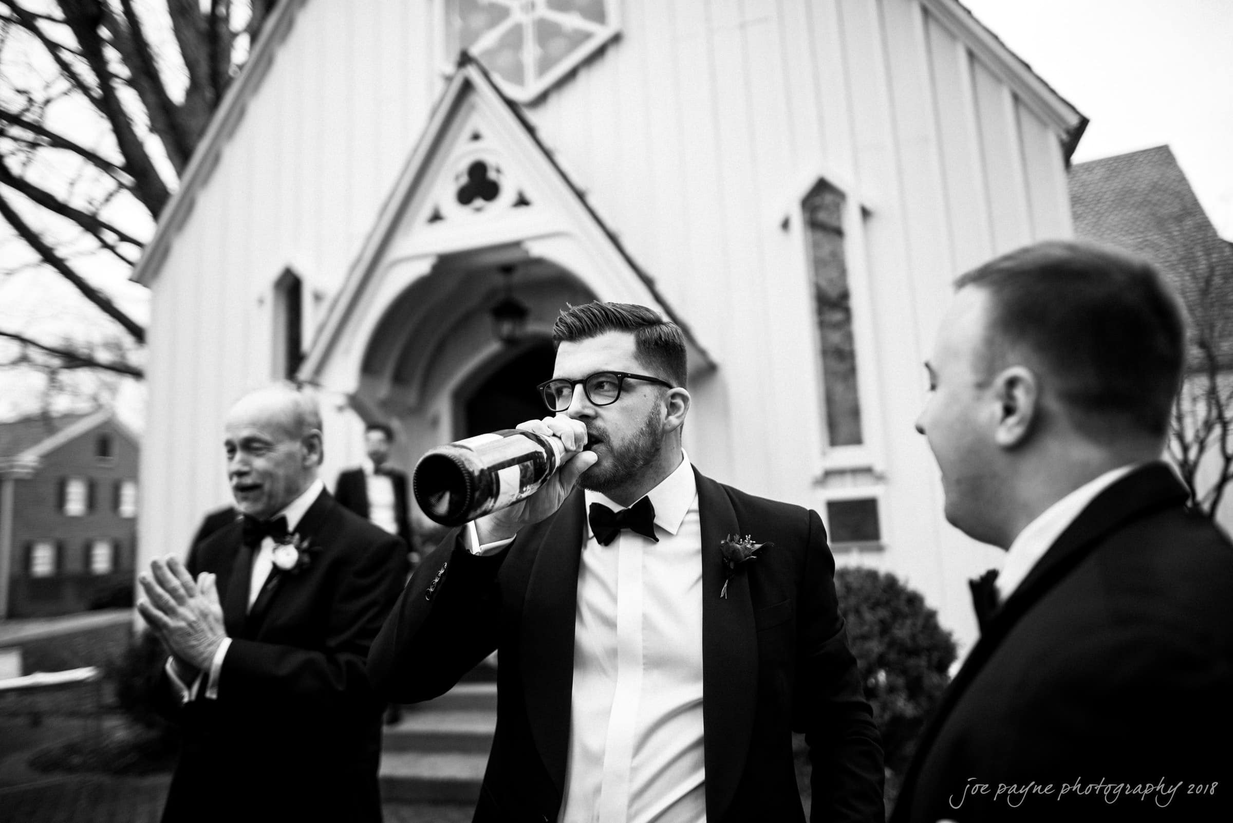 st. mary's & umstead wedding photographer - sarah & jake A man in a tuxedo drinks from a bottle outside a church, flanked by two other men in formal wear. The scene is in black and white, and the architecture of the church is visible in the background.