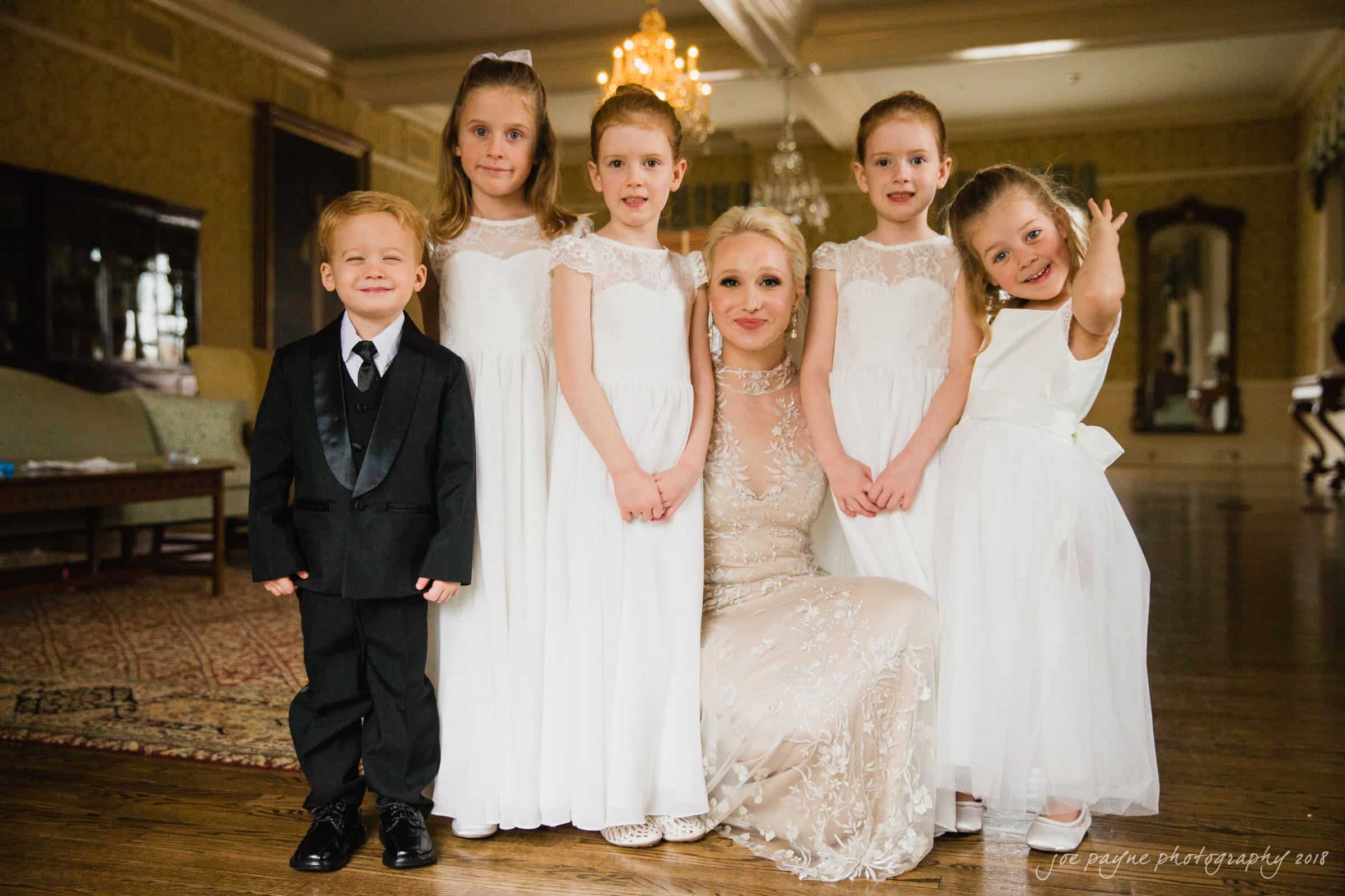 st. mary's & umstead wedding photographer - sarah & jake A woman in an elegant dress poses with five children wearing formal outfits. The children are dressed in white dresses and a black suit. They are all smiling and standing in a lavishly decorated room with chandeliers.