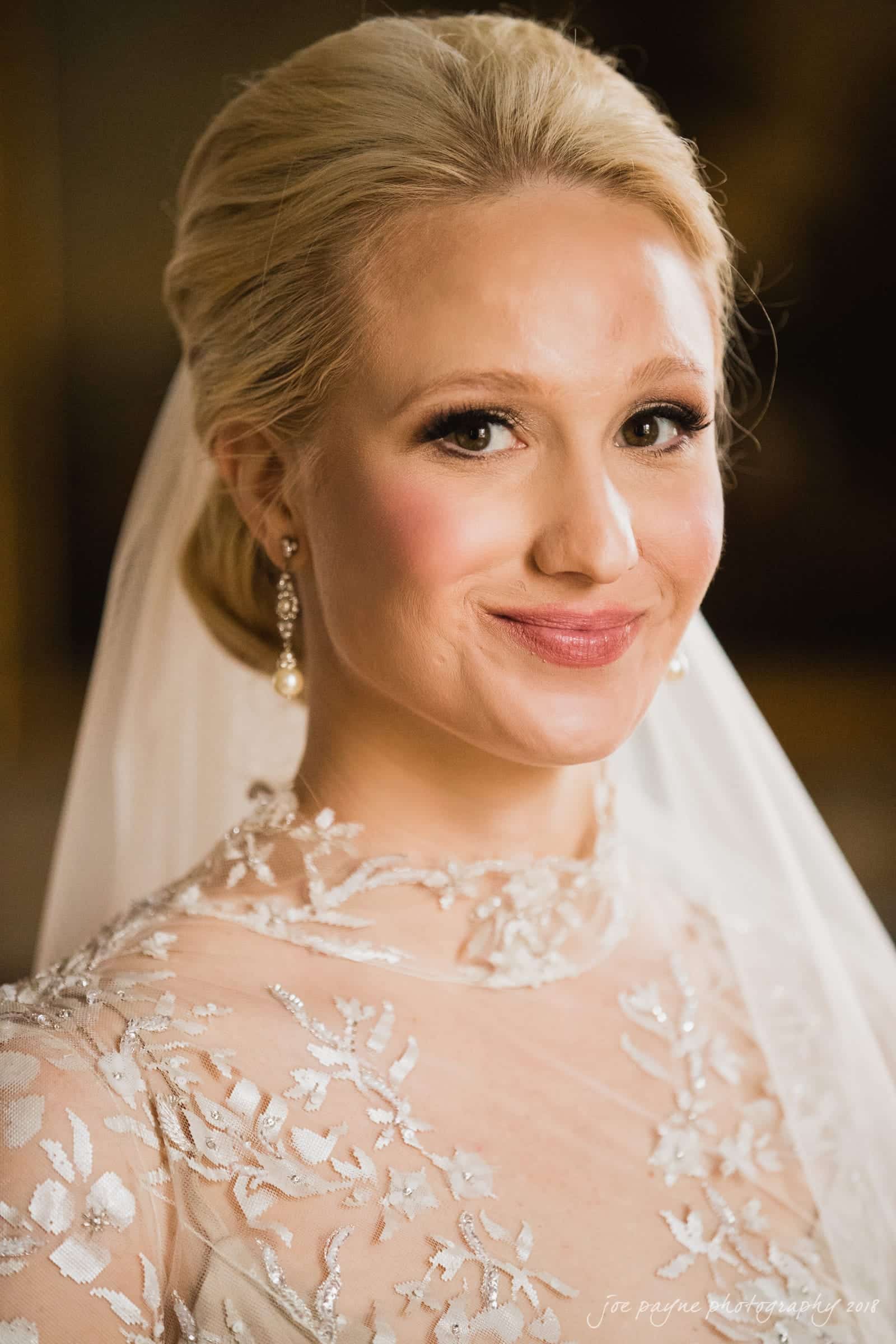 st. mary's & umstead wedding photographer - sarah & jake A bride with blonde hair smiles warmly at the camera. She wears a lace wedding dress and a veil. Her makeup is elegantly done, and she accessorizes with pearl earrings.