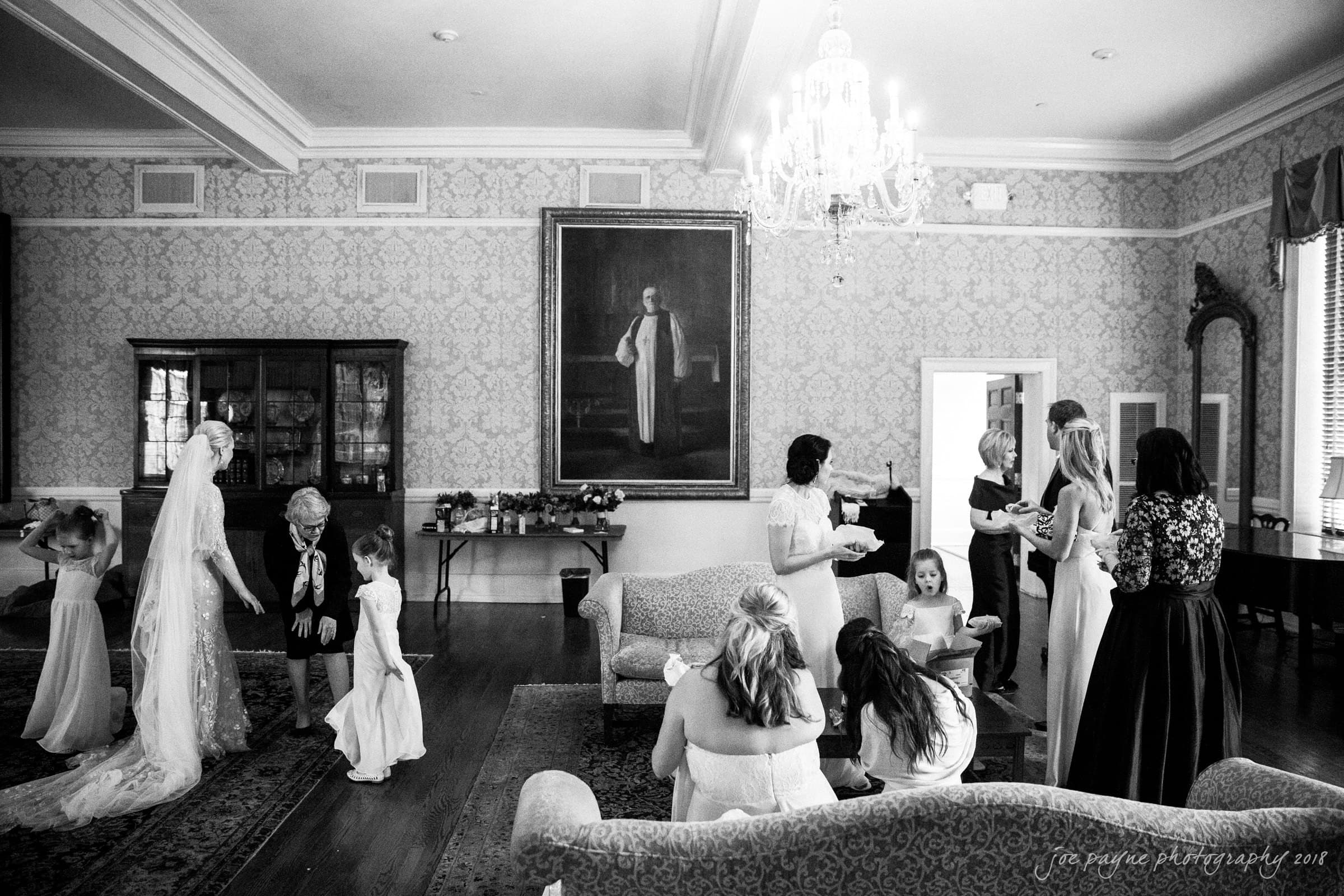 st. mary's & umstead wedding photographer - sarah & jake A black and white photo of a bridal party in an elegant room. Bridesmaids and flower girls are getting ready, while the bride stands with a long veil. A chandelier hangs from the ceiling, and a large portrait is on the wall. Luxurious sofas are in the room.