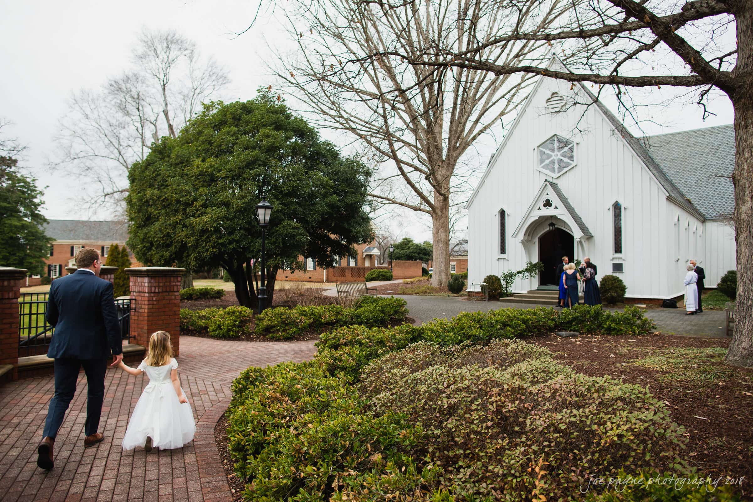 st. mary's & umstead wedding photographer - sarah & jake A man and a young girl in formal attire walk hand in hand along a brick path toward a white church. A group of people is gathered near the church entrance. Trees and greenery surround the area, and the sky is overcast.
