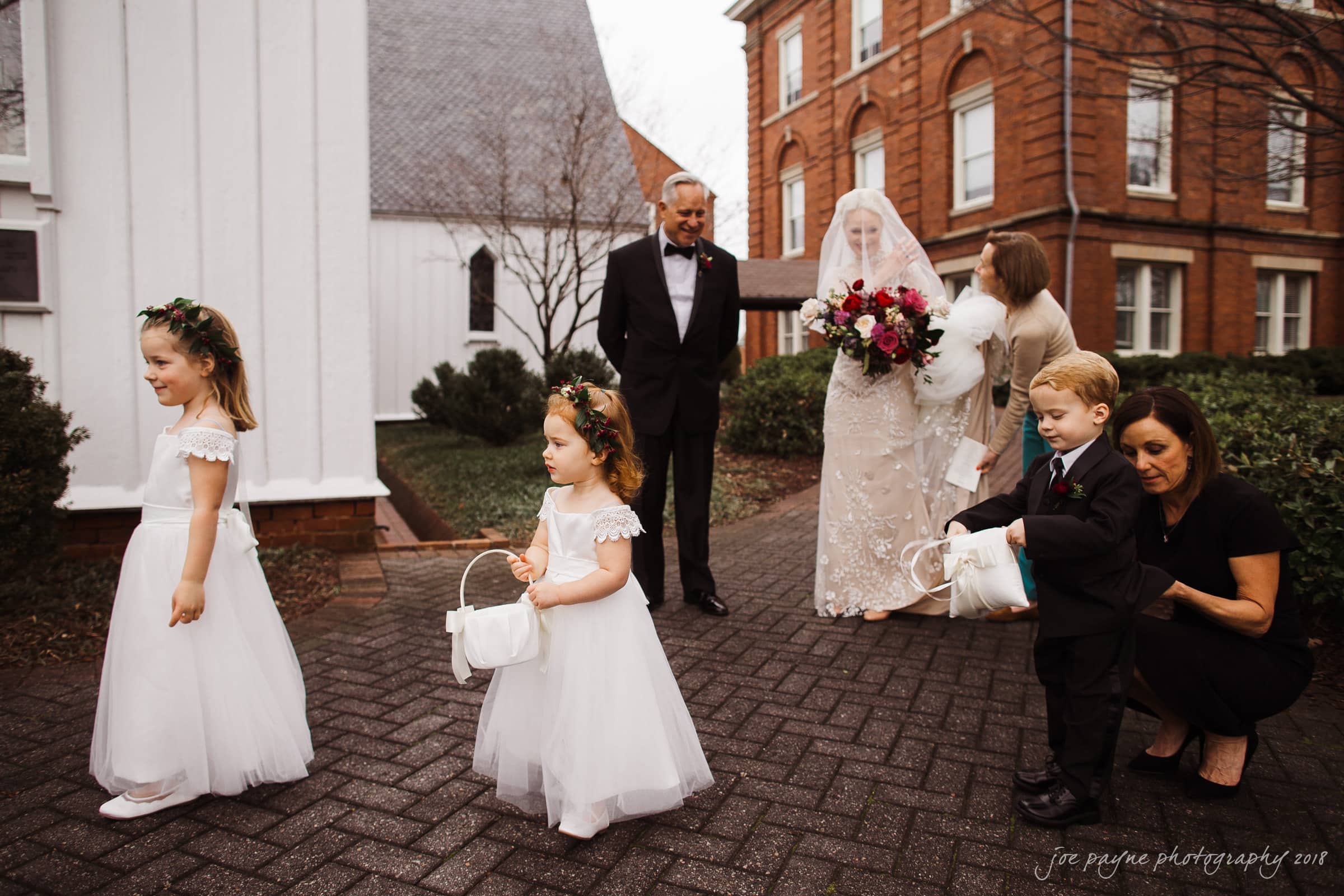 st. mary's & umstead wedding photographer - sarah & jake A bride in a white dress holds a bouquet, accompanied by two children in white dresses holding baskets. A man in a suit and a woman in a black outfit stand nearby. Theyre outside a church with brick buildings in the background.