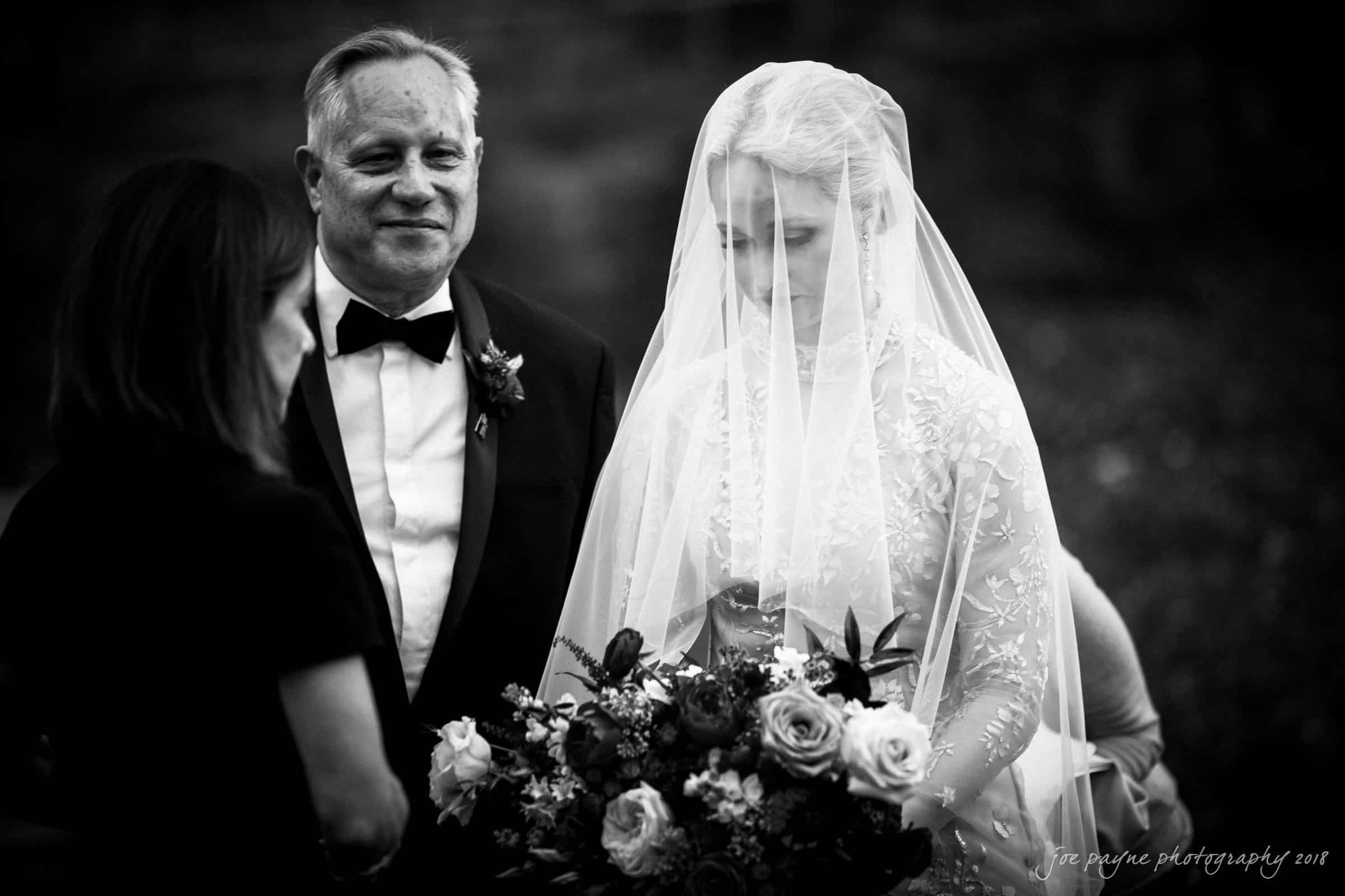 st. mary's & umstead wedding photographer - sarah & jake Bride in a lace wedding dress and veil stands holding a bouquet next to a man in a suit, while another woman turns toward them. The scene appears to be outdoors, possibly during a wedding ceremony, with a soft focus on the background.