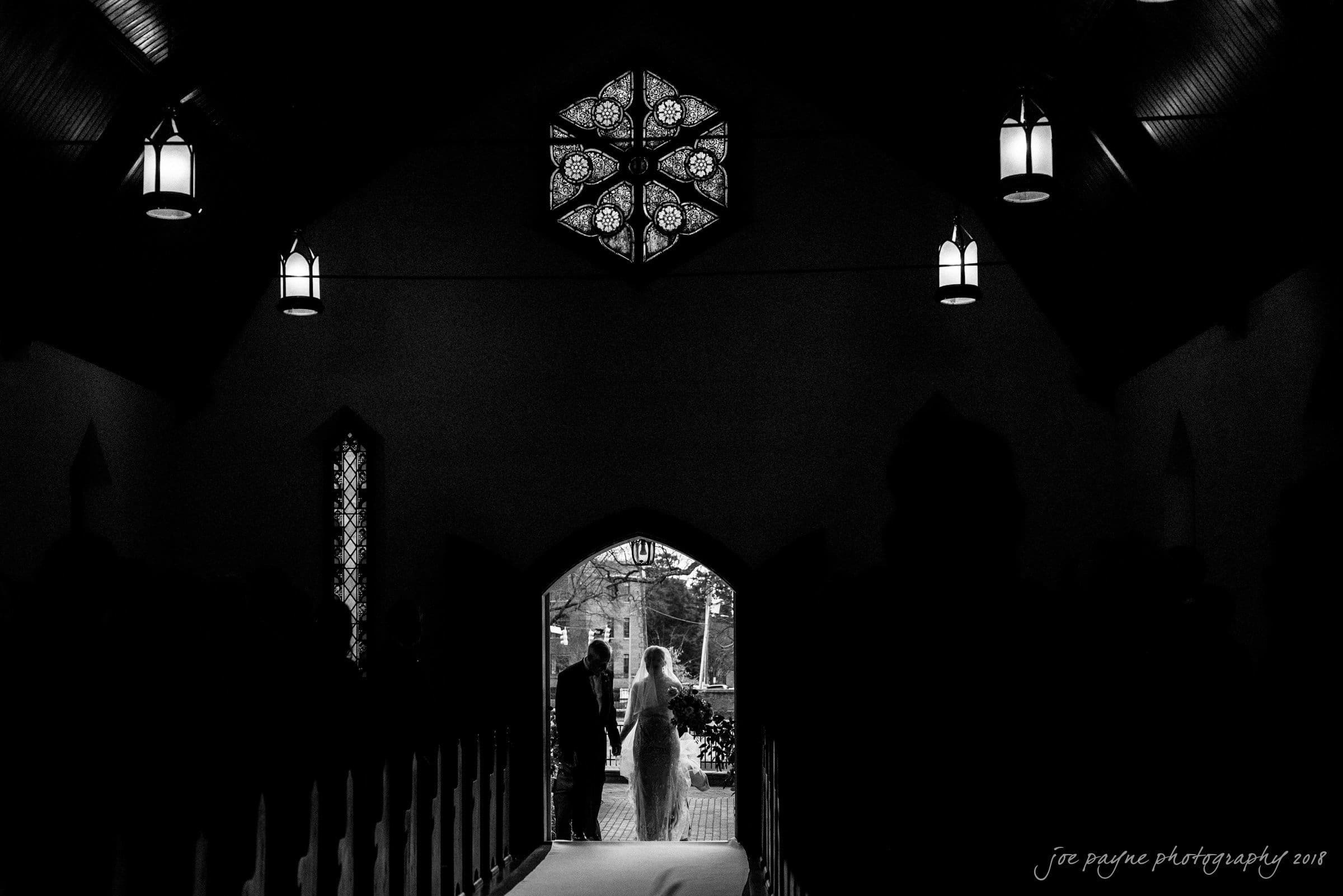 st. mary's & umstead wedding photographer - sarah & jake A bride and groom stand at the entrance of a dimly lit chapel, silhouetted against the light coming from outside. Stained glass and hanging lights are visible, creating a serene and intimate atmosphere.