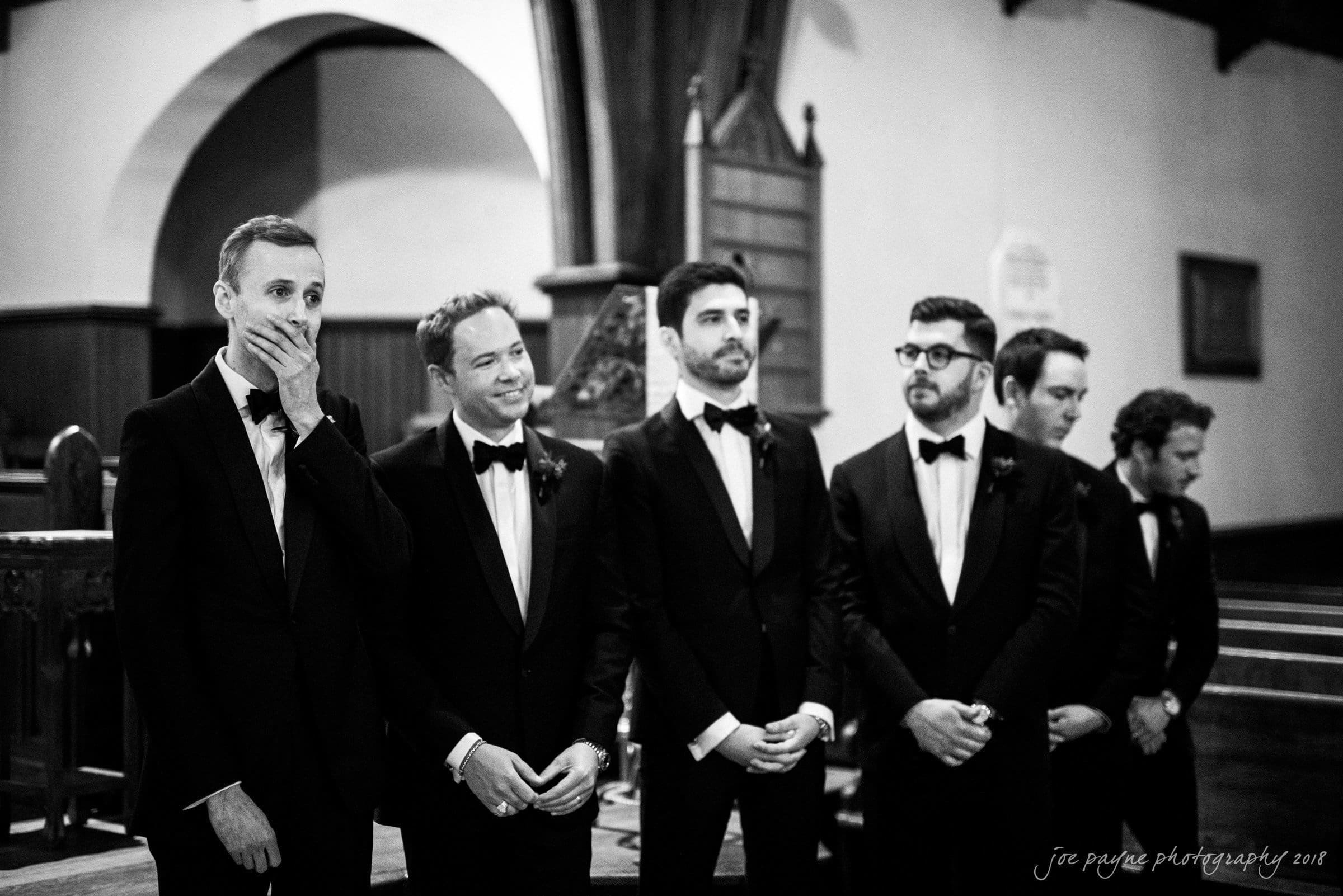 st. mary's & umstead wedding photographer - sarah & jake A group of men in tuxedos stand in a church, with one covering his mouth in anticipation. They appear to be part of a wedding party, waiting at the altar. The setting is elegant and formal.