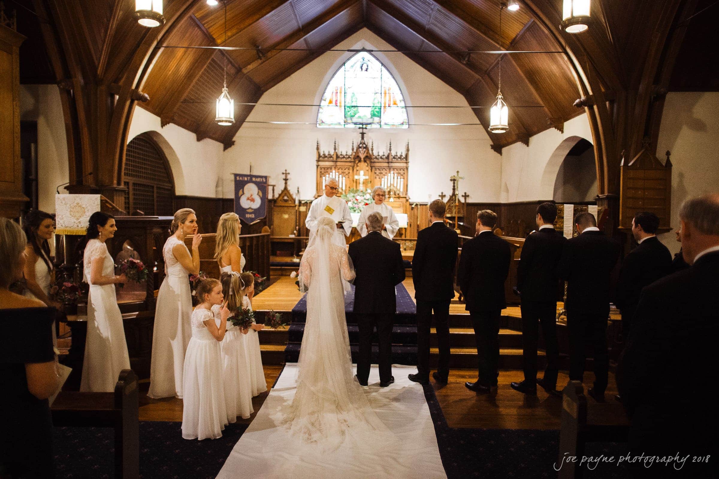 st. mary's & umstead wedding photographer - sarah & jake A bride and groom stand at the altar of a wooden church during their wedding ceremony. Bridesmaids and groomsmen are lined up on either side. A stained glass window is visible above the altar.