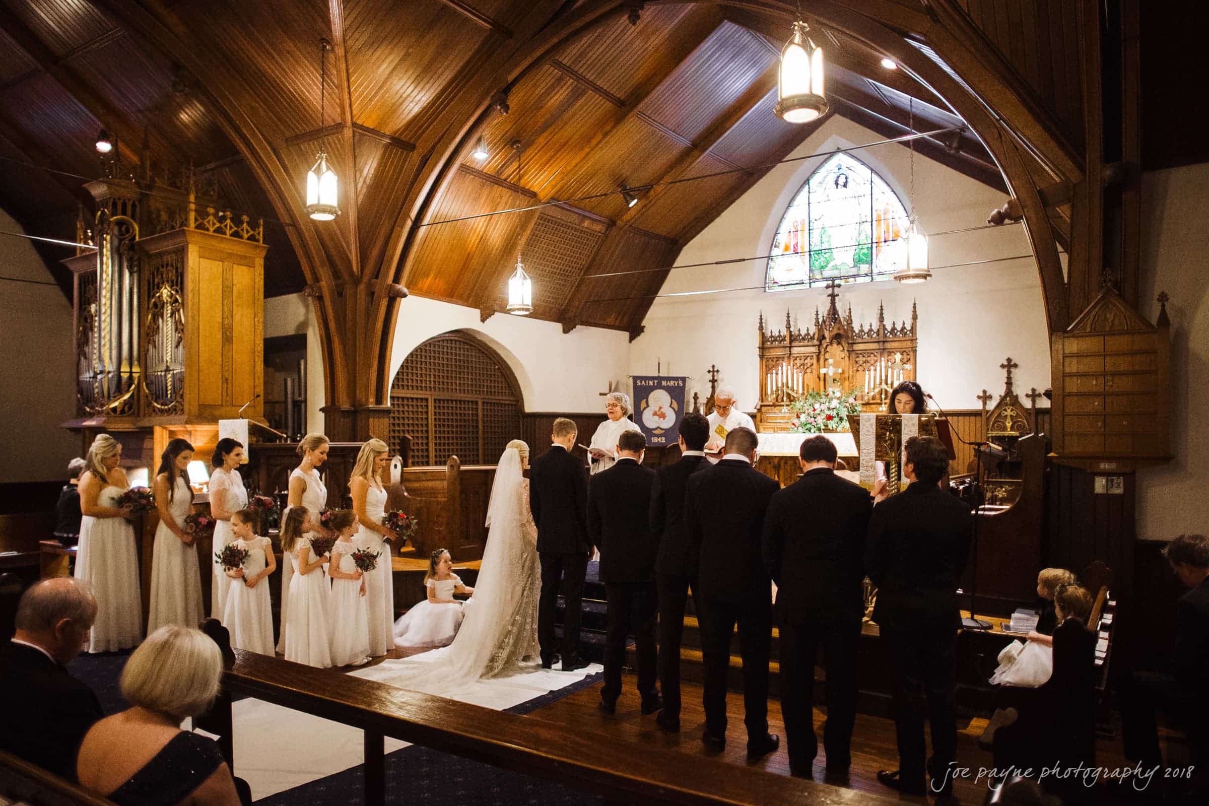 st. mary's & umstead wedding photographer - sarah & jake A bride and groom stand at the altar in a church, surrounded by their wedding party. The bridesmaids wear white dresses and hold bouquets, while the groomsmen wear black suits. The church has wooden beams and stained glass windows.