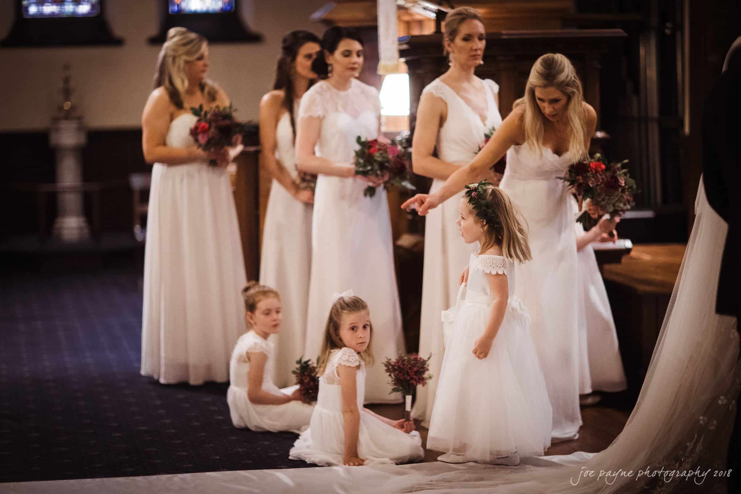st. mary's & umstead wedding photographer - sarah & jake A group of bridesmaids and flower girls in white dresses are gathered in a church. The bridesmaids hold bouquets, while the flower girls sit and stand nearby. The setting features stained glass windows and dim lighting.