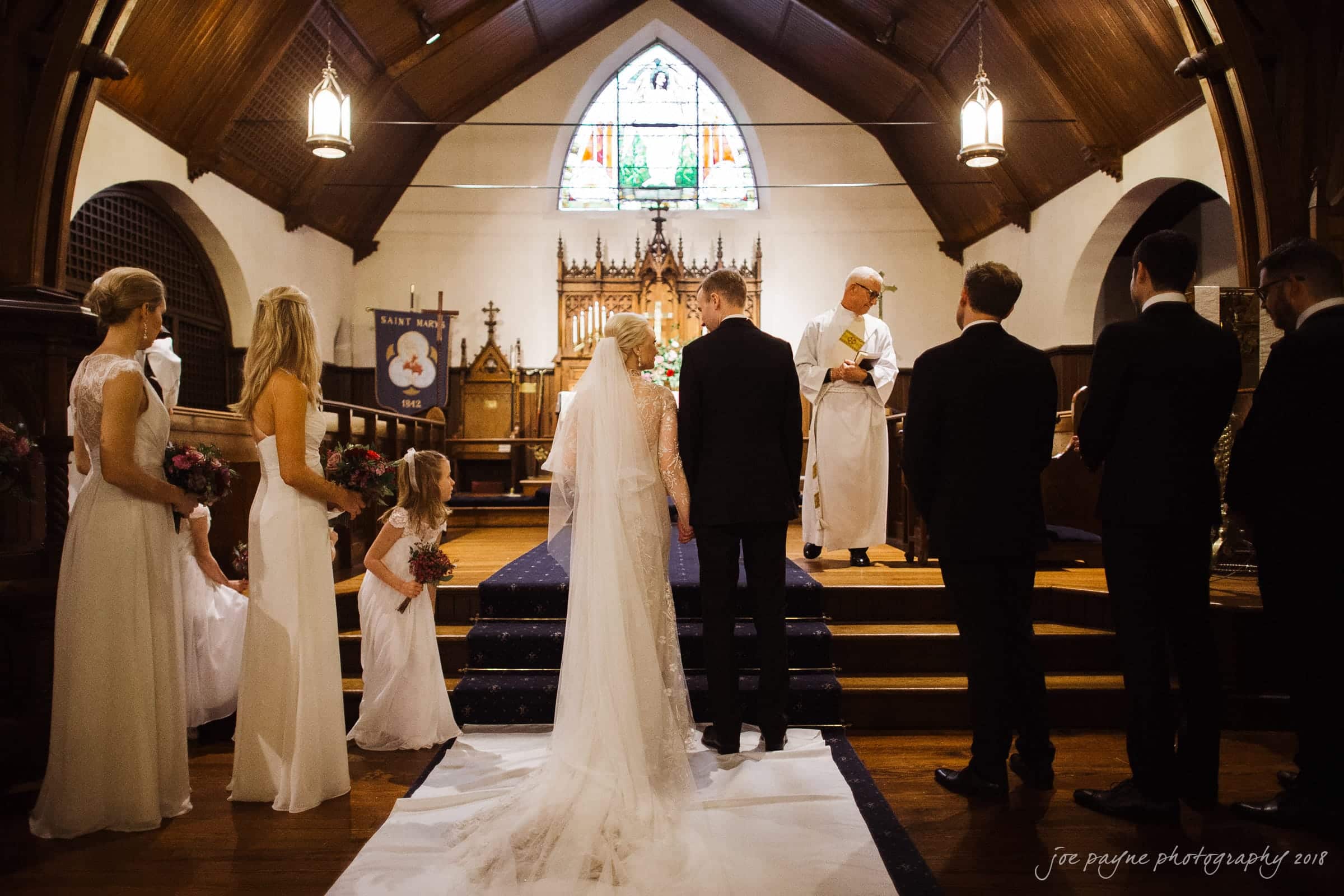 st. mary's & umstead wedding photographer - sarah & jake A wedding ceremony in a church, featuring a bride and groom standing before a priest. Bridesmaids and a flower girl in white dresses stand on the left, while groomsmen in black suits stand on the right. A stained glass window is visible in the background.
