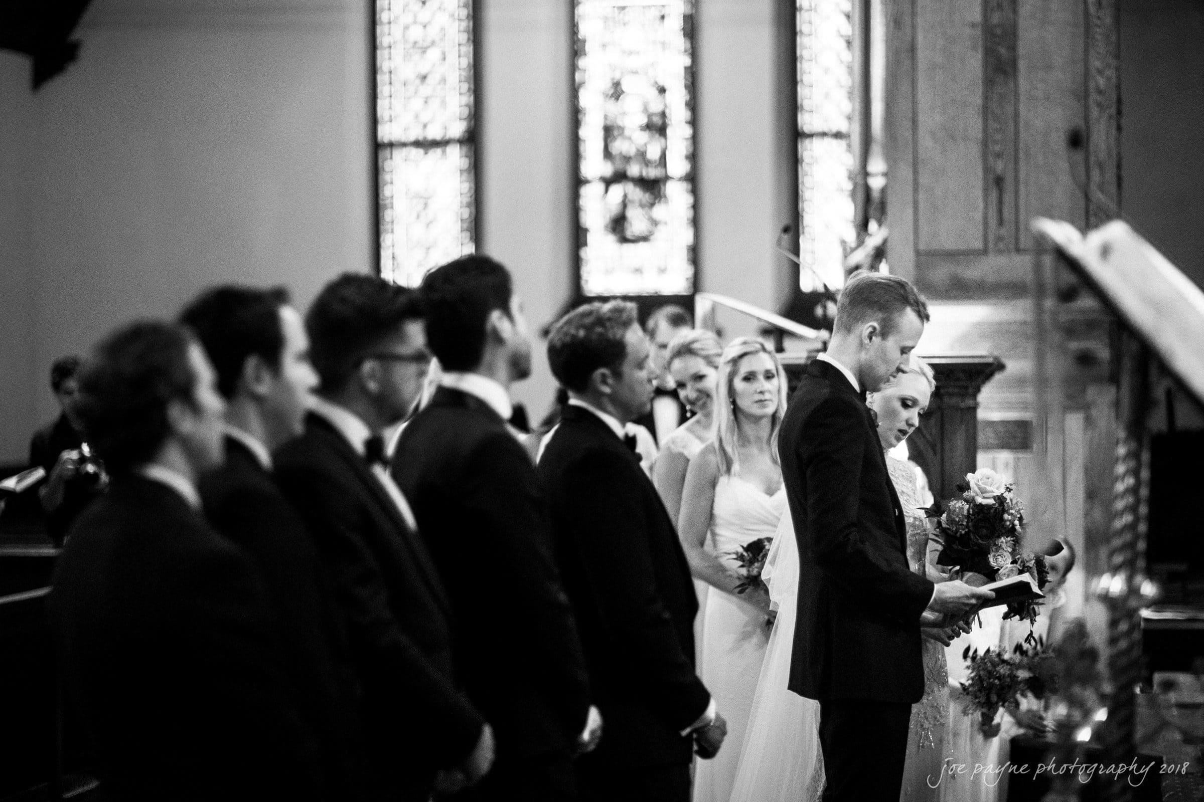 st. mary's & umstead wedding photographer - sarah & jake A black-and-white photo of a wedding ceremony in a church. The groom, wearing a suit, stands holding a book and reading. The bride, in a white dress, stands beside him with a bouquet. The stained glass windows and guests are visible in the background.