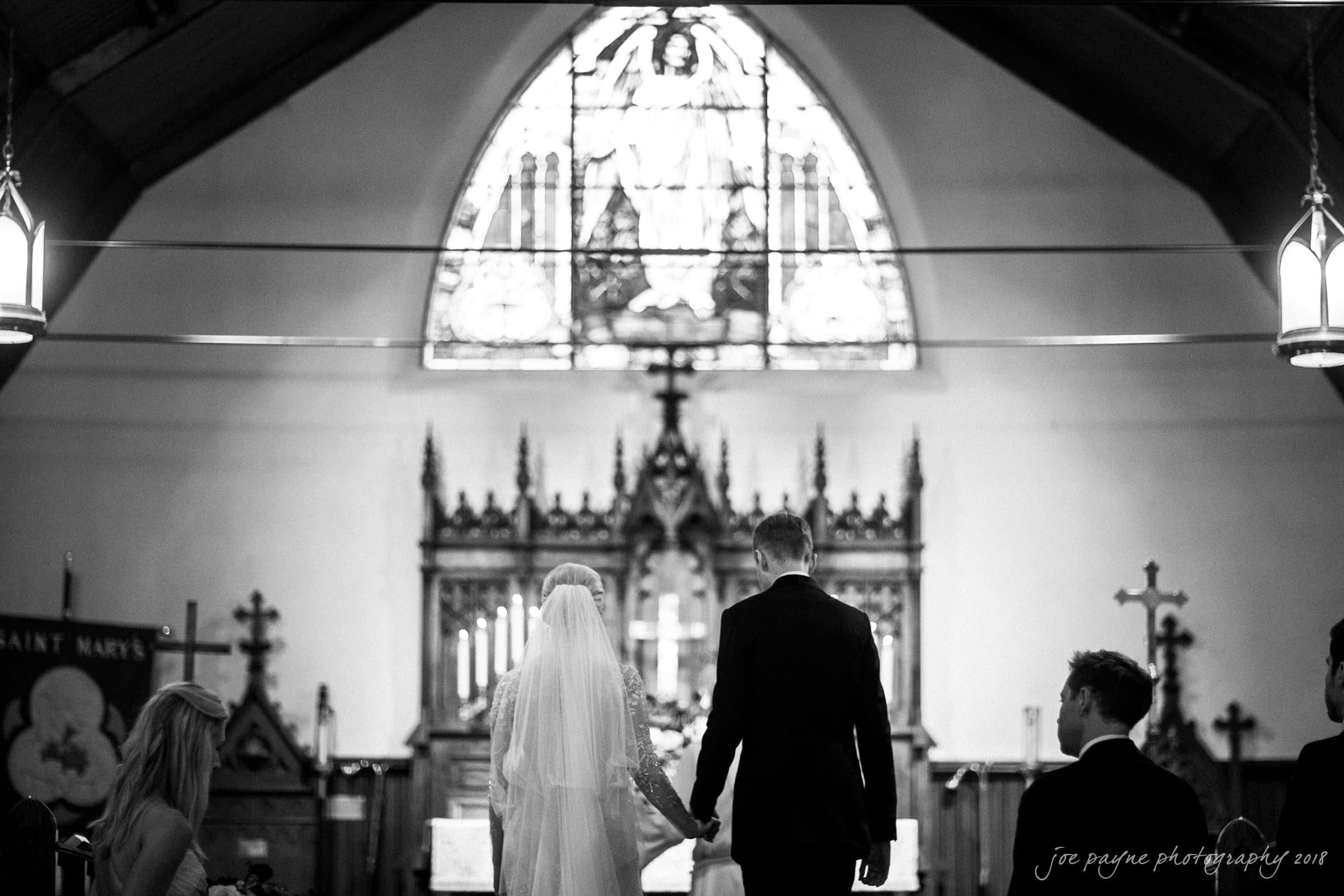 st. mary's & umstead wedding photographer - sarah & jake A bride and groom stand at the altar in a church, holding hands. The setting features stained glass windows and wood paneling. Two people are seated nearby, observing the ceremony. The image is in black and white.