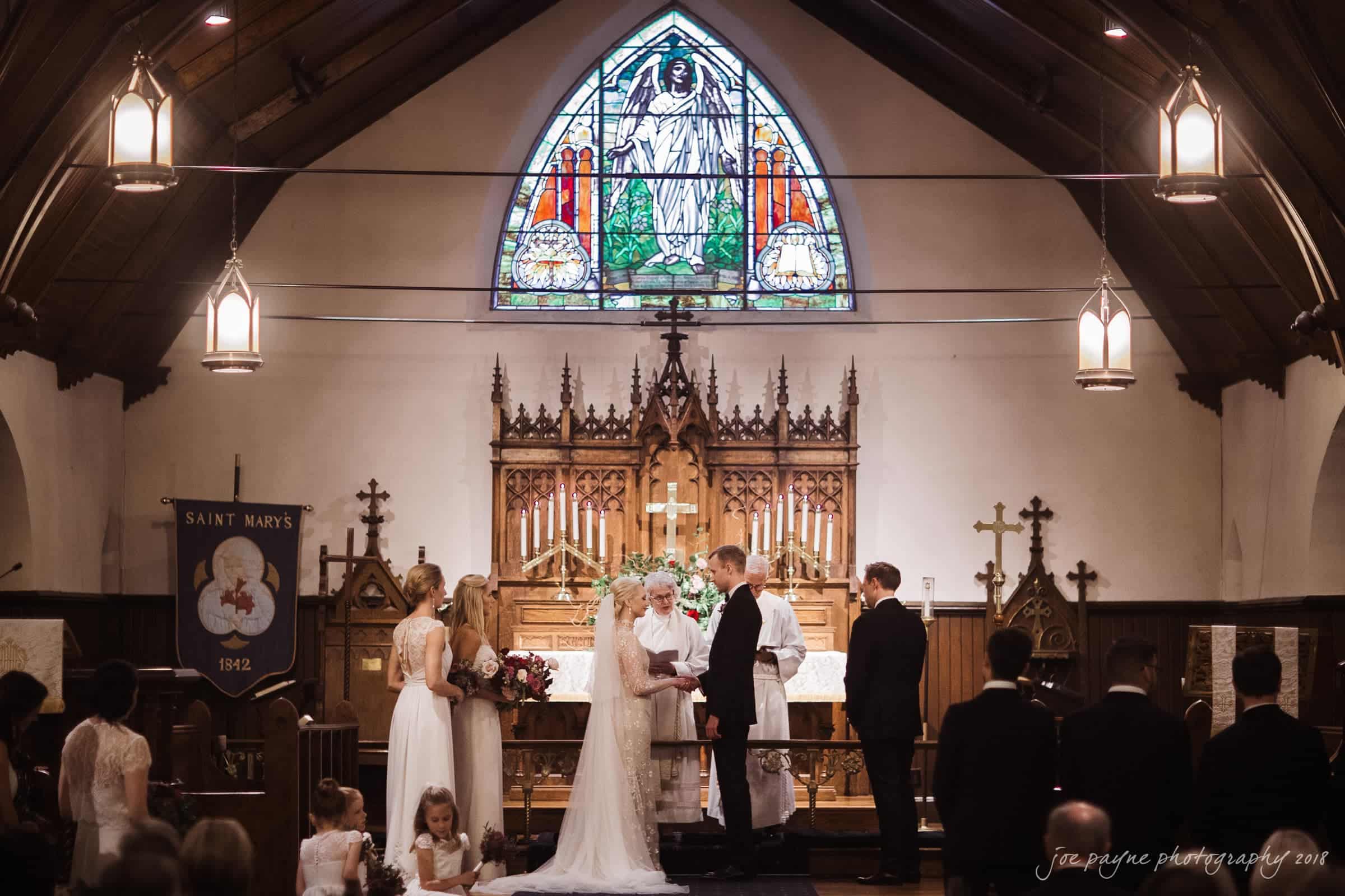 st. mary's & umstead wedding photographer - sarah & jake A wedding ceremony taking place in a church. The bride and groom stand at the altar, surrounded by the wedding party. A stained glass window is visible above them, and guests are seated in the pews.