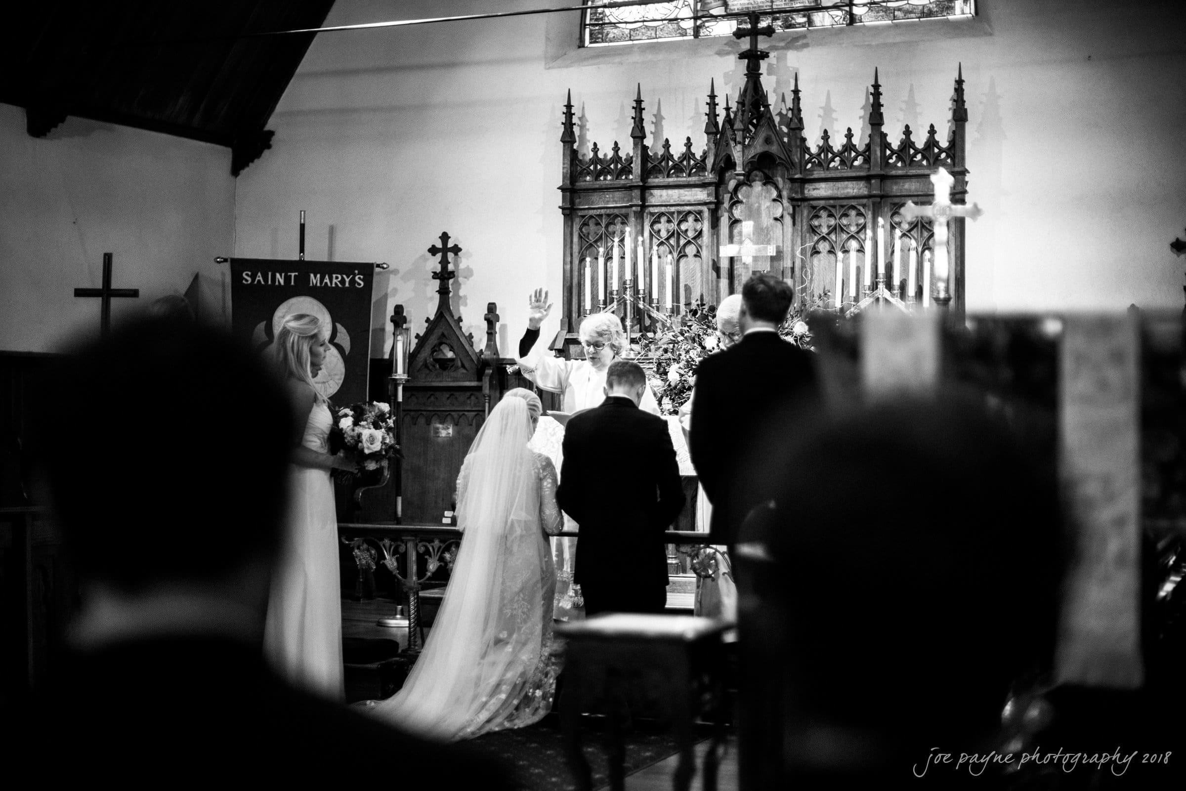 st. mary's & umstead wedding photographer - sarah & jake A black-and-white photo of a wedding ceremony in a church. The bride and groom kneel before a priest who is raising his hand in blessing. Two bridesmaids stand beside them. An ornate altar with candles is in the background.