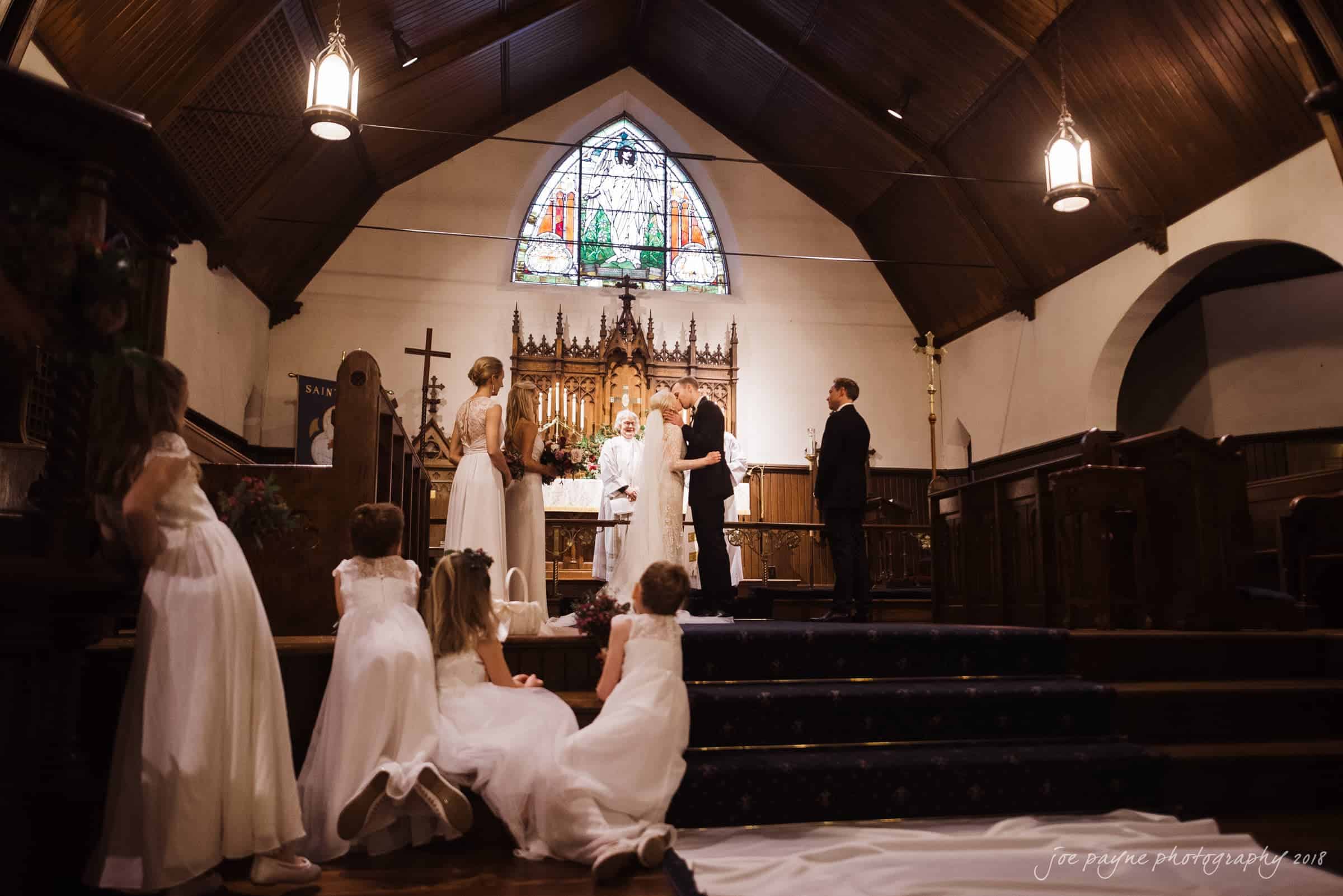 st. mary's & umstead wedding photographer - sarah & jake A wedding ceremony in a church with a stained glass window. The bride and groom stand at the altar with a minister. Bridesmaids in white dresses kneel nearby, arranging the brides long train on the wooden floor.
