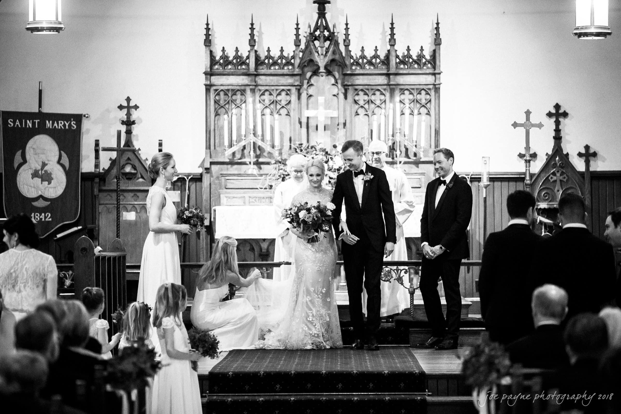 st. mary's & umstead wedding photographer - sarah & jake A black and white photo of a wedding ceremony in a church. The bride and groom stand at the altar holding hands. Bridesmaids and groomsmen are on either side. Floral arrangements and a banner with a cross are visible in the background.