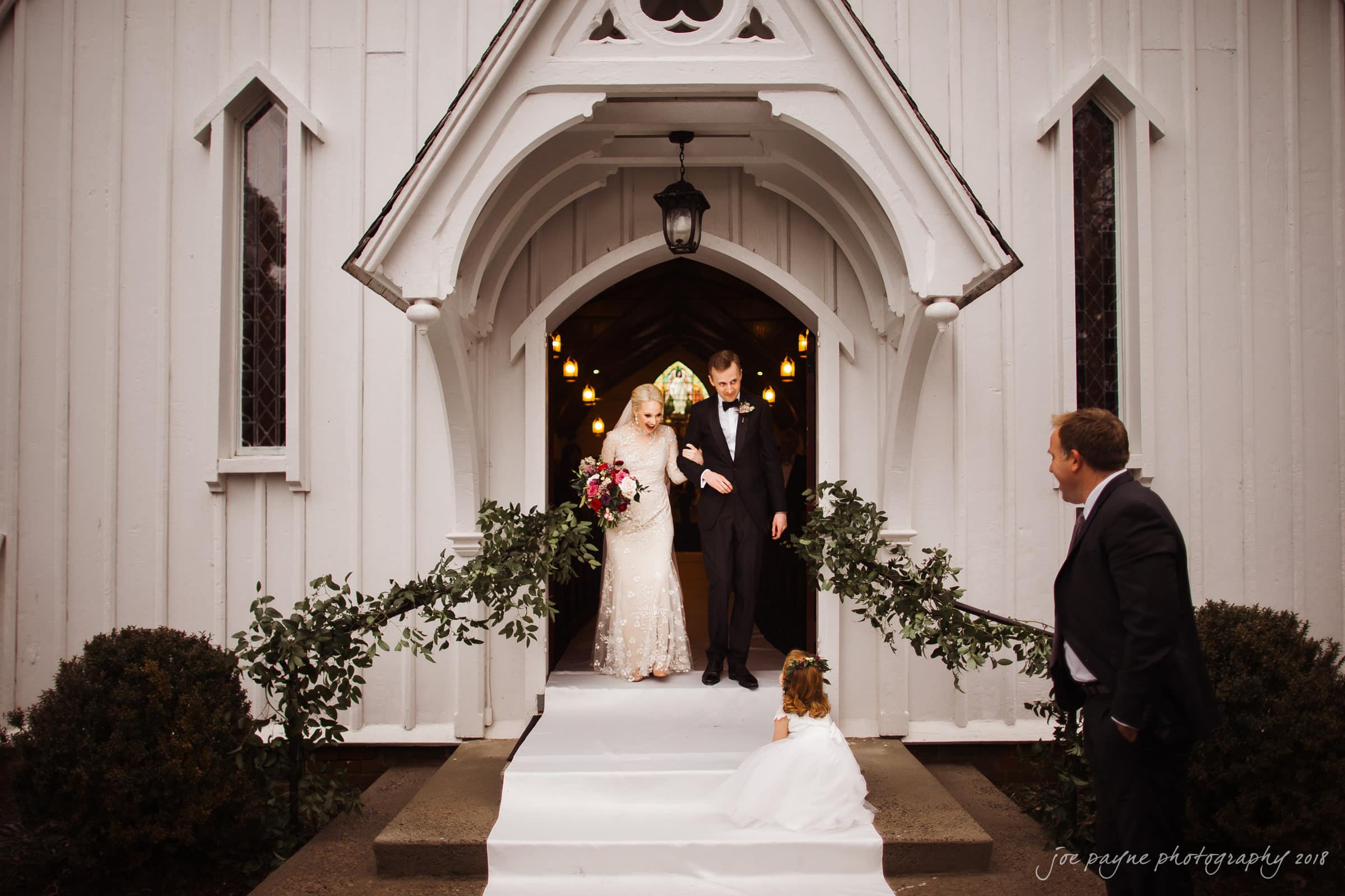st. mary's & umstead wedding photographer - sarah & jake Bride and groom exiting a white wooden church, walking down a decorated staircase. The bride holds a bouquet and wears a lace gown, while the groom is in a black suit. A flower girl sits on the steps, and a man stands nearby.