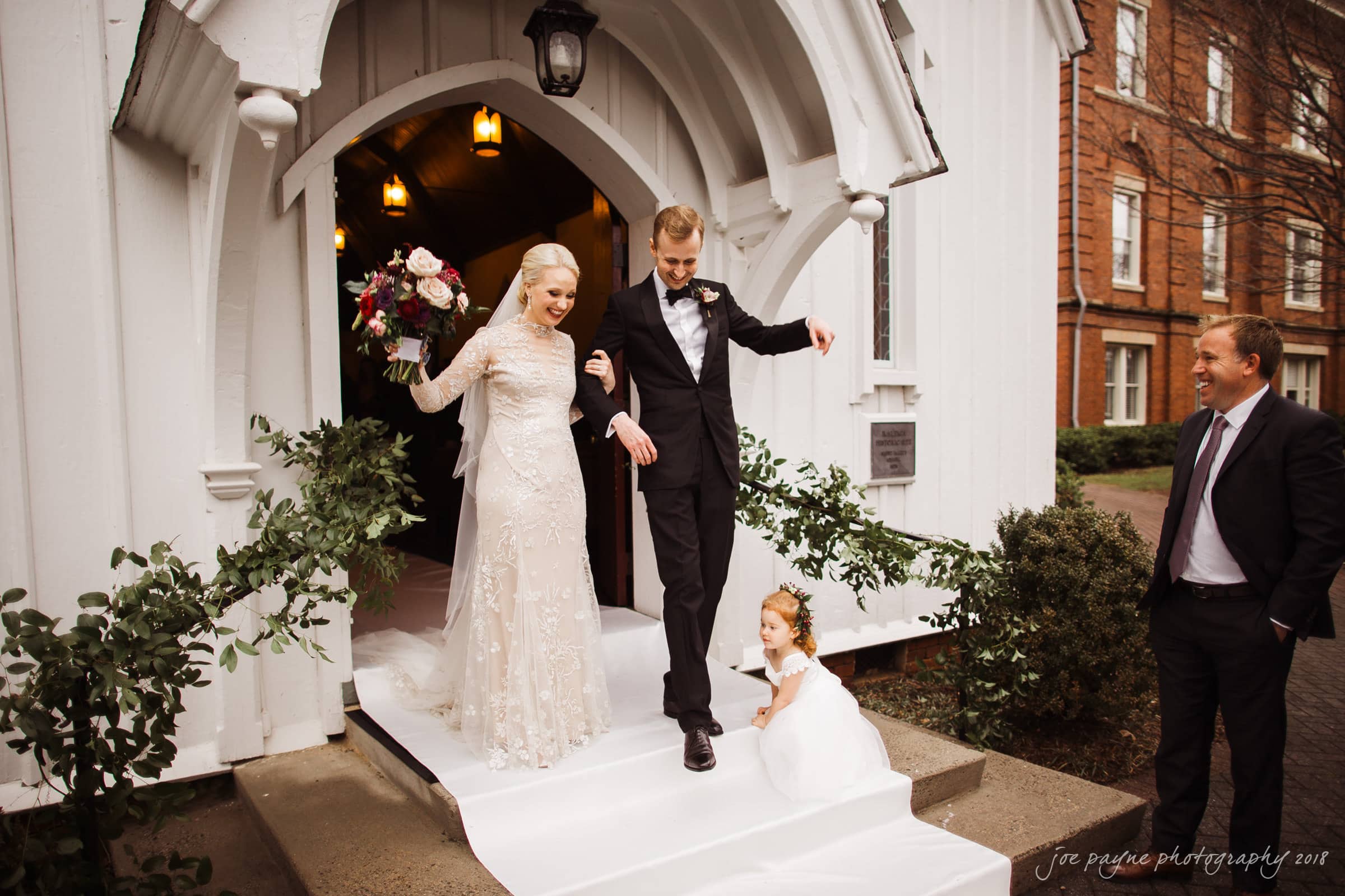 st. mary's & umstead wedding photographer - sarah & jake A bride and groom happily exit a white chapel, holding hands. The bride wears a long white dress, and a small child stands in front with a basket. A man in a suit watches them from the side. Theres greenery around the entrance.