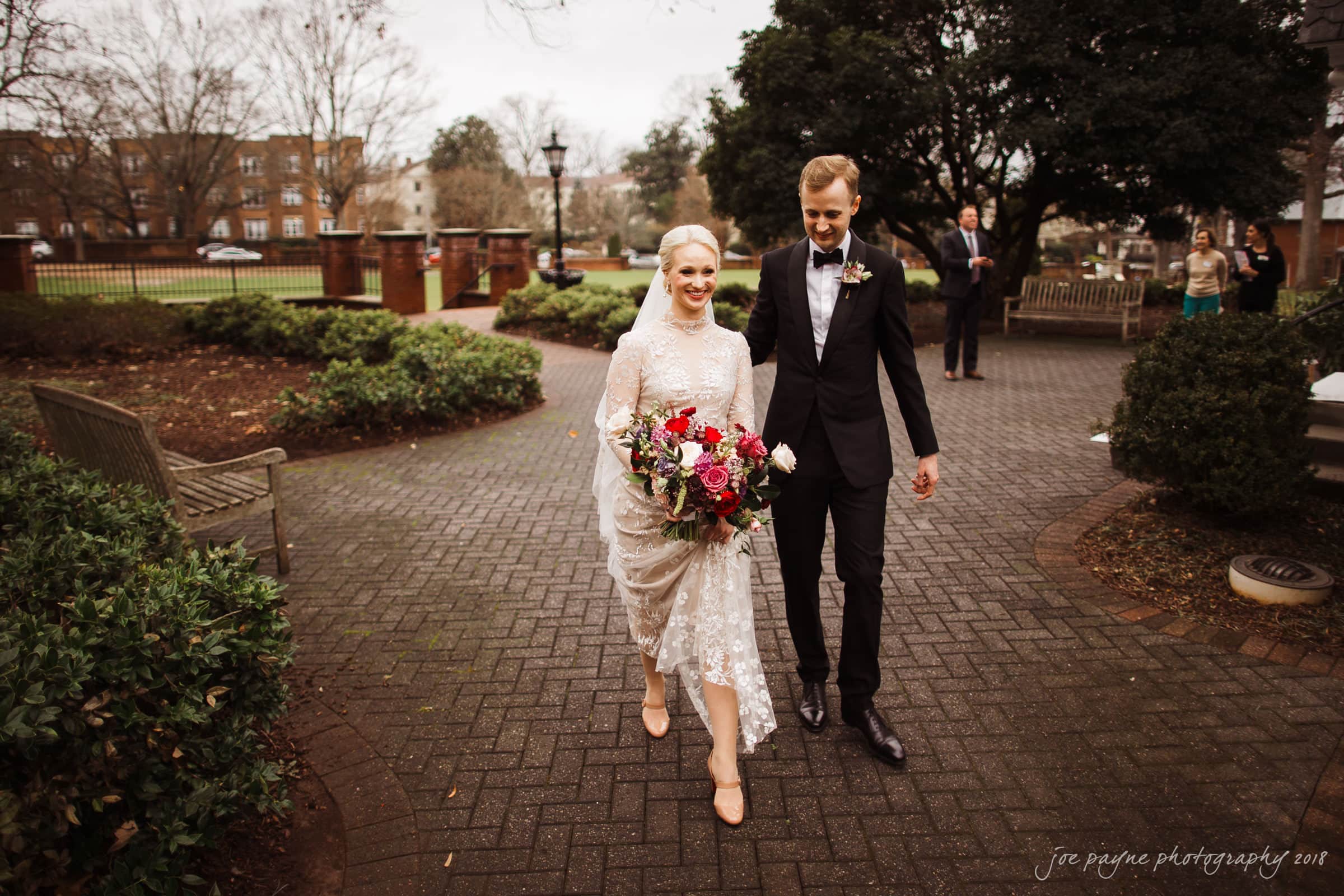 st. mary's & umstead wedding photographer - sarah & jake A bride and groom are walking outside on a brick path. The bride is wearing a white lace dress and holding a bouquet of colorful flowers. The groom is in a black suit. Trees and bushes surround the area. A few people are in the background.